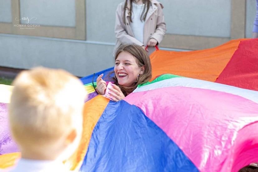 Uśmiechnięta animatorka wewnątrz kolorowej chusty animacyjnej, z widocznym fragmentem dziecka w lewym dolnym rogu i osobą dorosłą w tle, podczas animacji dla dzieci na świeżym powietrzu.
