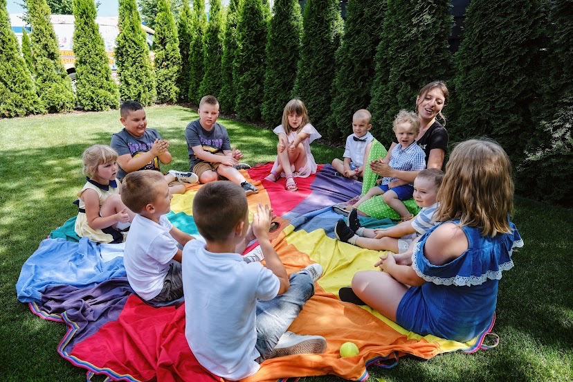 Grupa dzieci siedzi na kolorowej chuście na trawie podczas zajęć z animatorką, w tle rząd wysokich tuj.