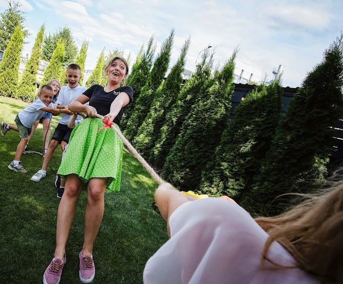 Grupa dzieci i dorosła kobieta w zielonej spódnicy grają w przeciąganie liny na trawniku, tło stanowią wysokie tuje.