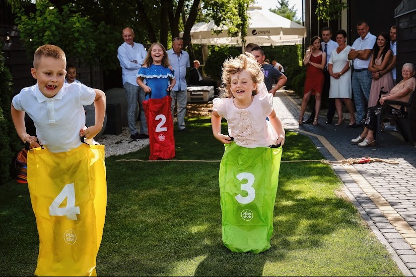 Radosne dzieci uczestniczą w wyścigu w workach podczas imprezy plenerowej, z widocznymi workami oznaczonymi numerami 2, 3 i 4, a w tle obserwujący dorośli i dzieci.