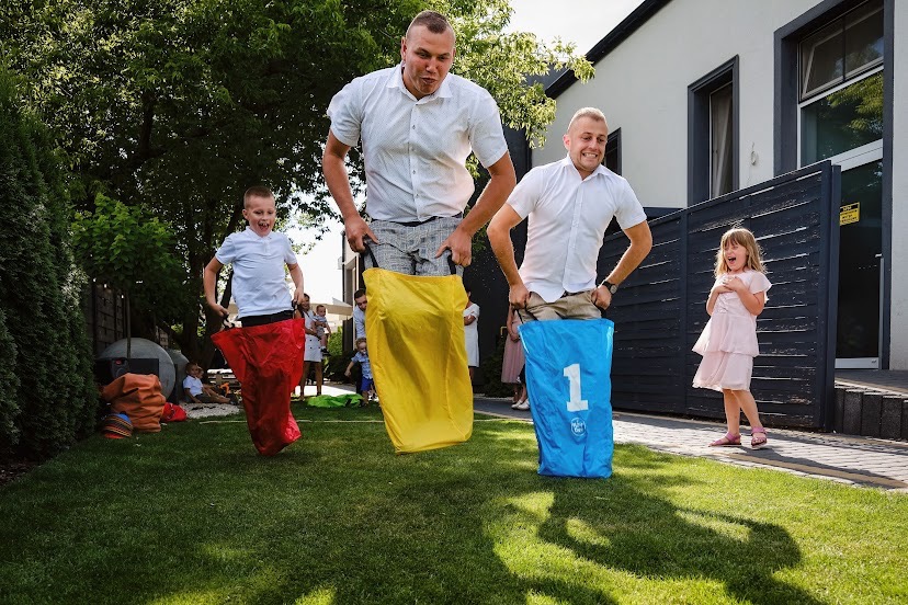 Zawody w skokach w workach podczas imprezy plenerowej: chłopiec w czerwonym worku, mężczyzna w żółtym, mężczyzna w niebieskim z numerem 1, na tle zielonego trawnika, domu i obserwujących dzieci...
