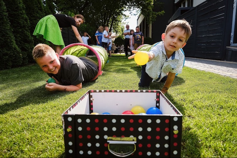 Chłopcy czołgają się przez tunele na trawie, celując piłkami do pudełka w kropki podczas zabawy z animatorem w ogrodzie.