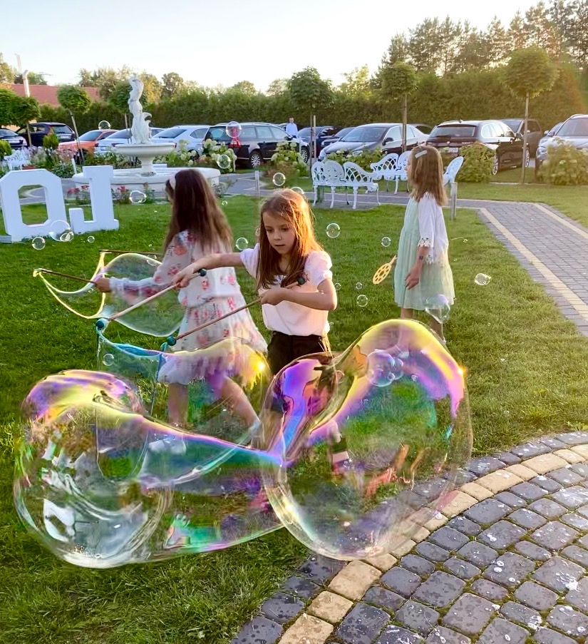 Trzy dziewczynki w Sochaczewie bawią się na trawniku, tworząc ogromne bańki mydlane za pomocą specjalnych kijków i sznurków. W tle widać fontannę, białe dekoracje i zaparkowane samochody.