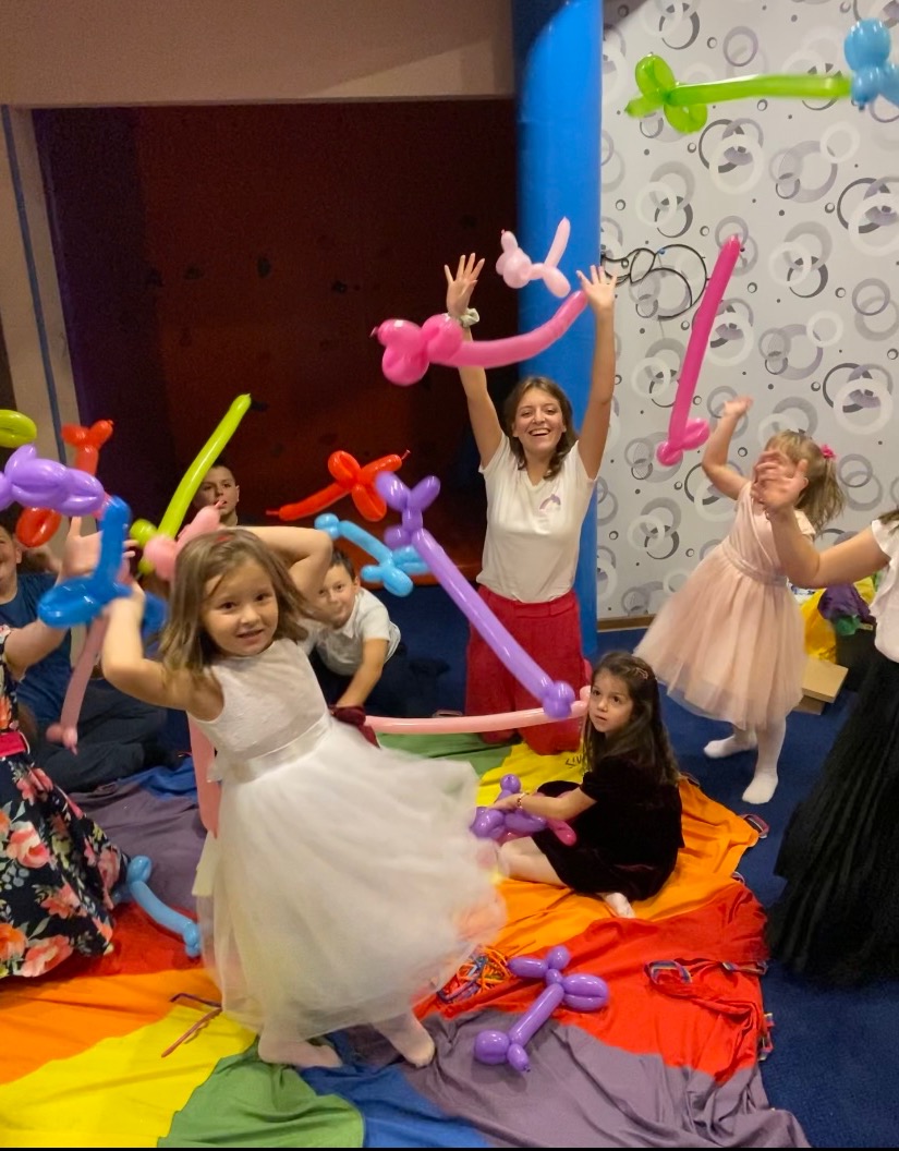 Dzieci w sukienkach bawią się balonami modelowanymi w kształty zwierząt, pod nadzorem animatorki na kolorowej macie w sali zabaw.