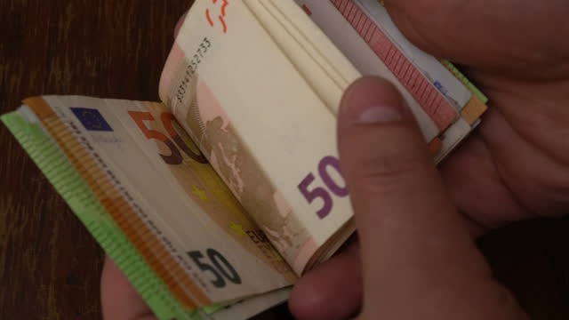 Dłoń przewraca plik banknotów 50 euro na drewnianym stole, widoczny fragment numeru seryjnego.