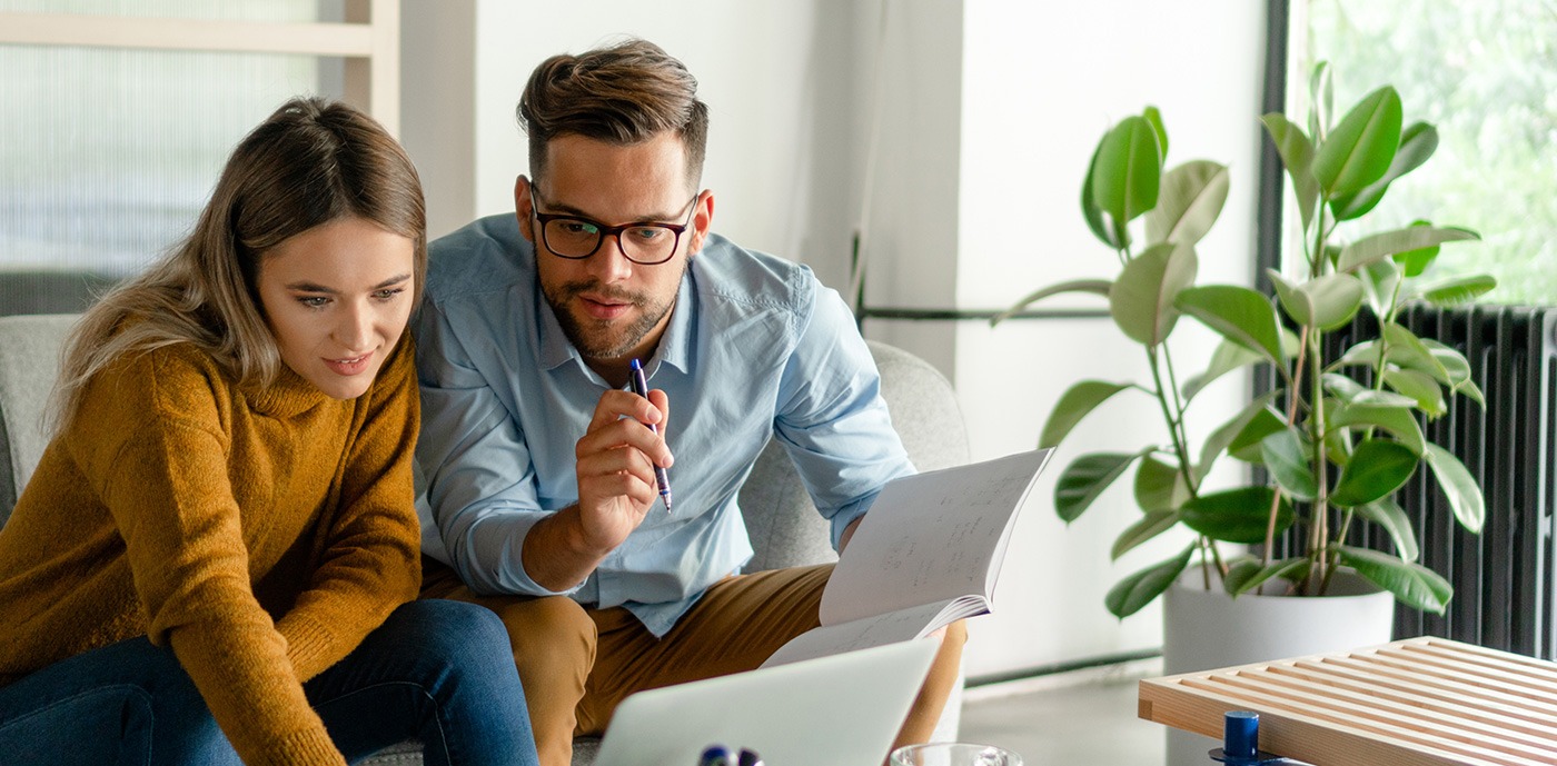 Para siedząca na kanapie analizuje dokumenty i dane na laptopie, mężczyzna trzyma długopis w dłoni, obok stoi roślina doniczkowa.