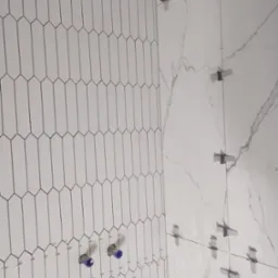 Montaż białych, podłużnych płytek w kształcie migdałów na ścianie, z widocznymi niebieskimi zaślepkami na rurach i systemem poziomowania płytek.