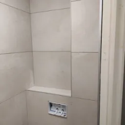 Ściana łazienki wyłożona beżowymi płytkami ceramicznymi, wnęka z półką i zamontowany stelaż podtynkowy WC, widoczne notatki na płytkach i ościeżnicy.