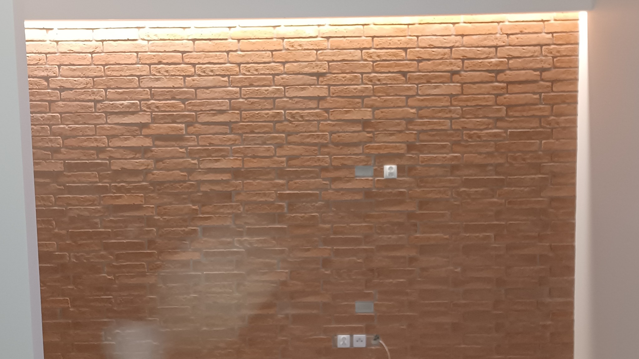 Ściana wyłożona cegłą dekoracyjną w pomieszczeniu, widoczne gniazdka elektryczne i włączniki światła, oświetlenie górne.
