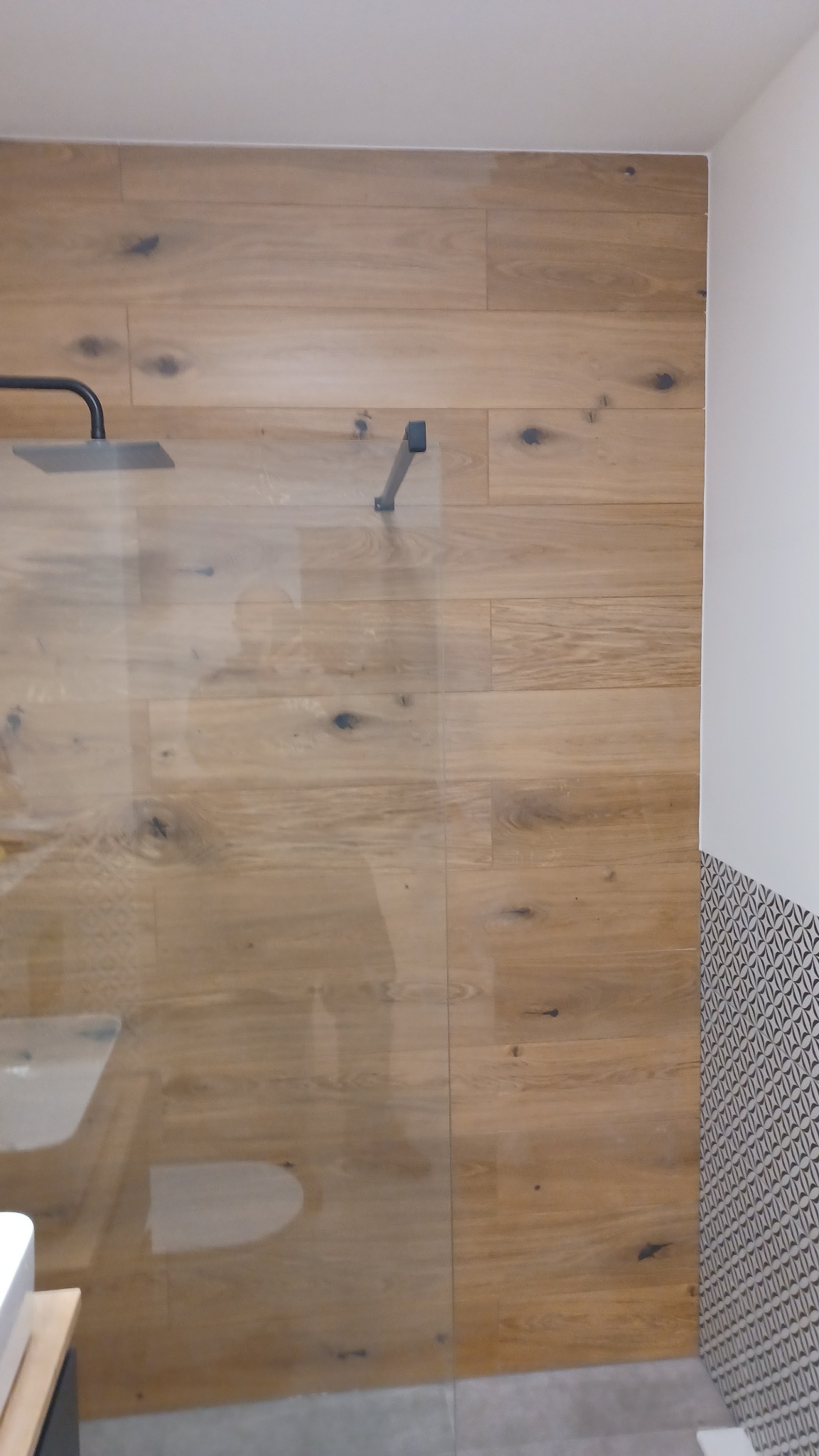 Kabina prysznicowa ze ścianami wyłożonymi panelami imitującymi drewno, czarny deszczownica i mocowanie szyby, fragment dekoracyjnej mozaiki na ścianie obok.