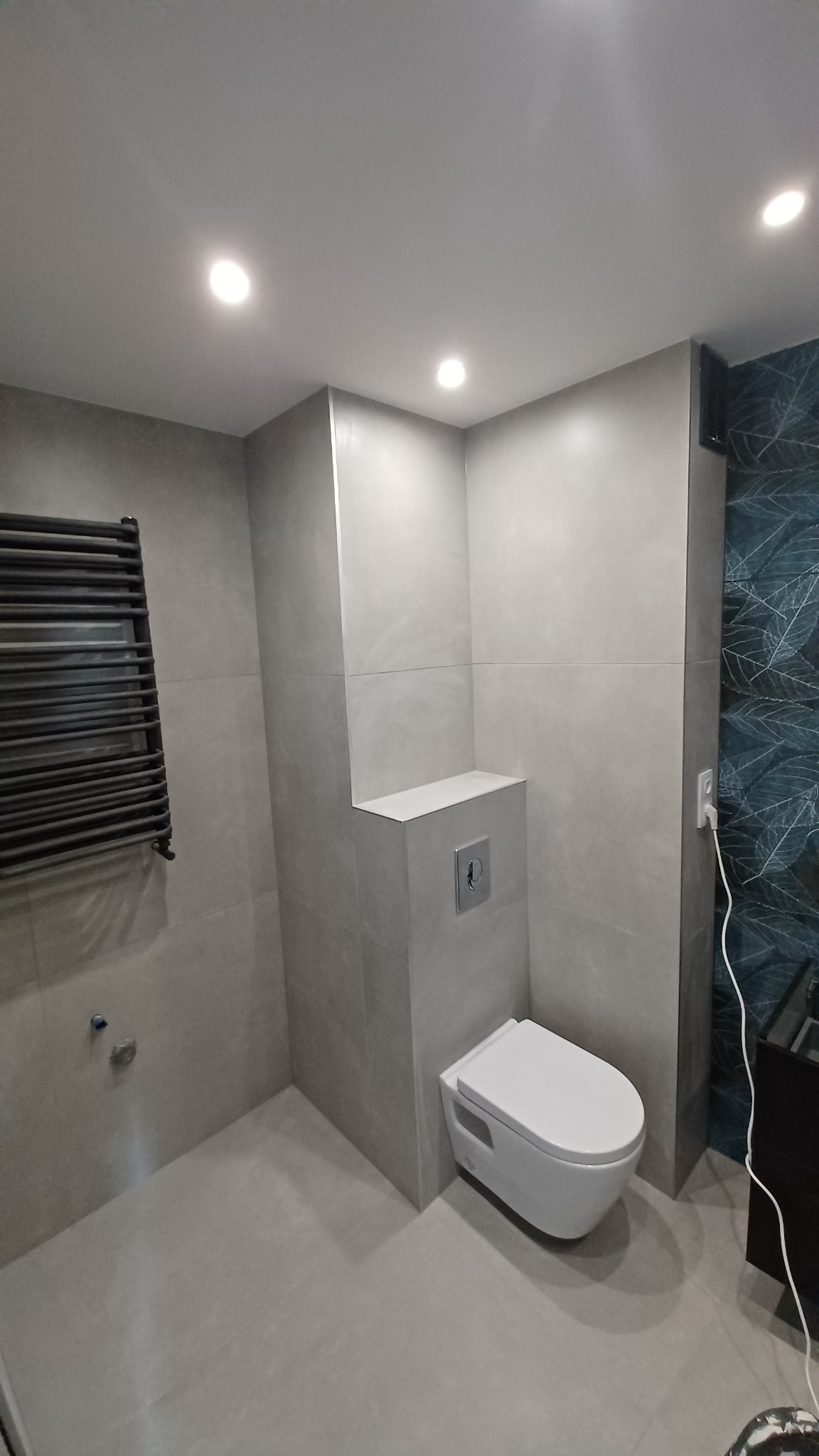 Nowoczesna łazienka w szarościach z podwieszaną toaletą i grzejnikiem drabinkowym, akcentem jest ściana z motywem liści, oświetlenie punktowe w suficie.