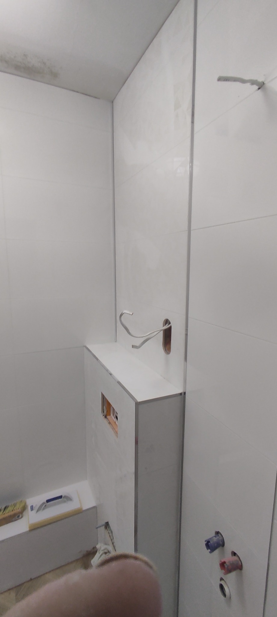 Łazienka w trakcie wykańczania: białe płytki na ścianach, zabudowa stelaża WC z otworem rewizyjnym, wystające kable elektryczne, narzędzia glazurnicze w tle, widoczne przyłącza wodne.