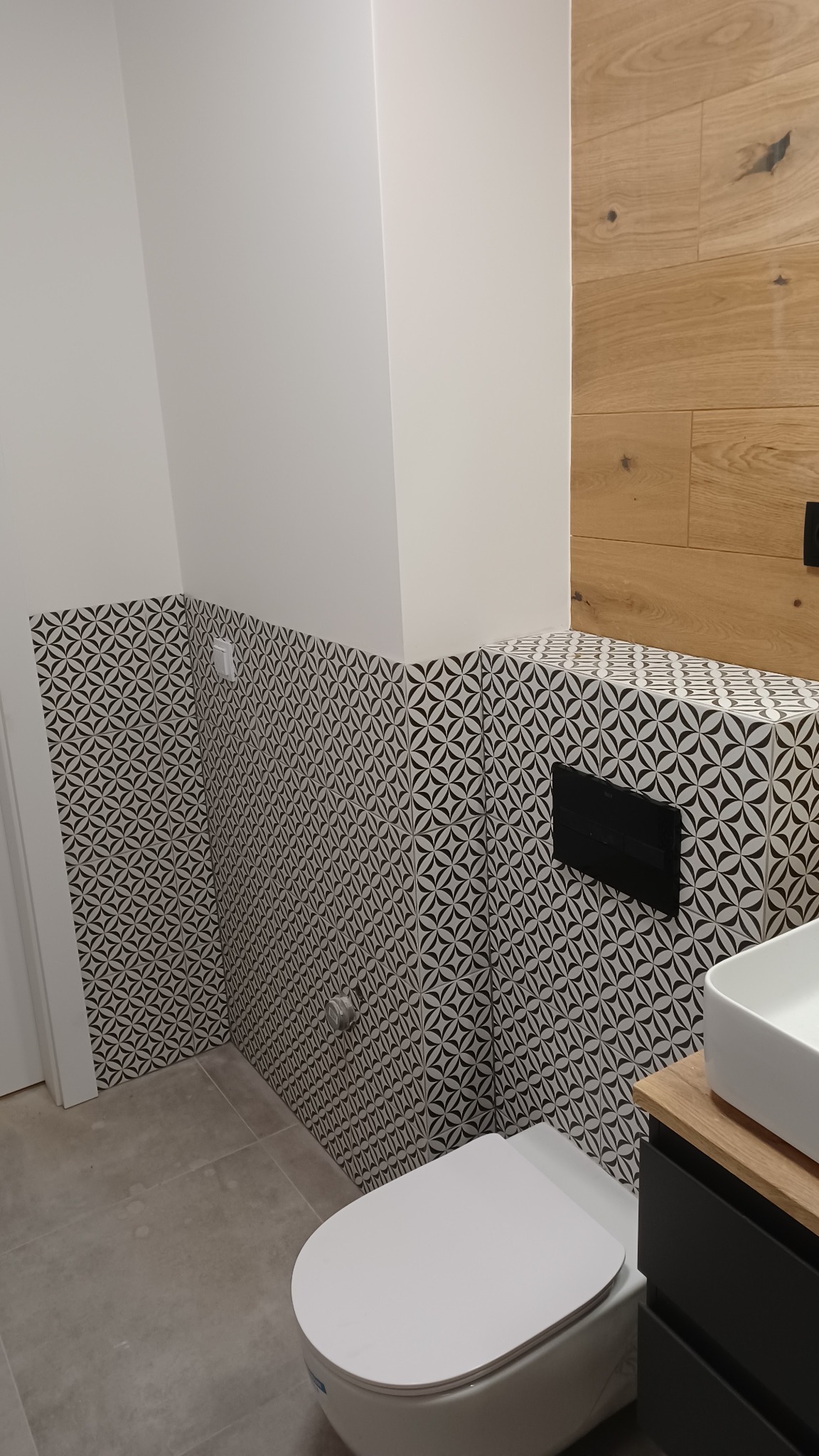 Nowoczesna łazienka z białą miską WC i wzorzystymi, czarno-białymi płytkami na ścianach, uzupełniona drewnianą okładziną ściany i czarną armaturą, widoczne szare płytki podłogowe.