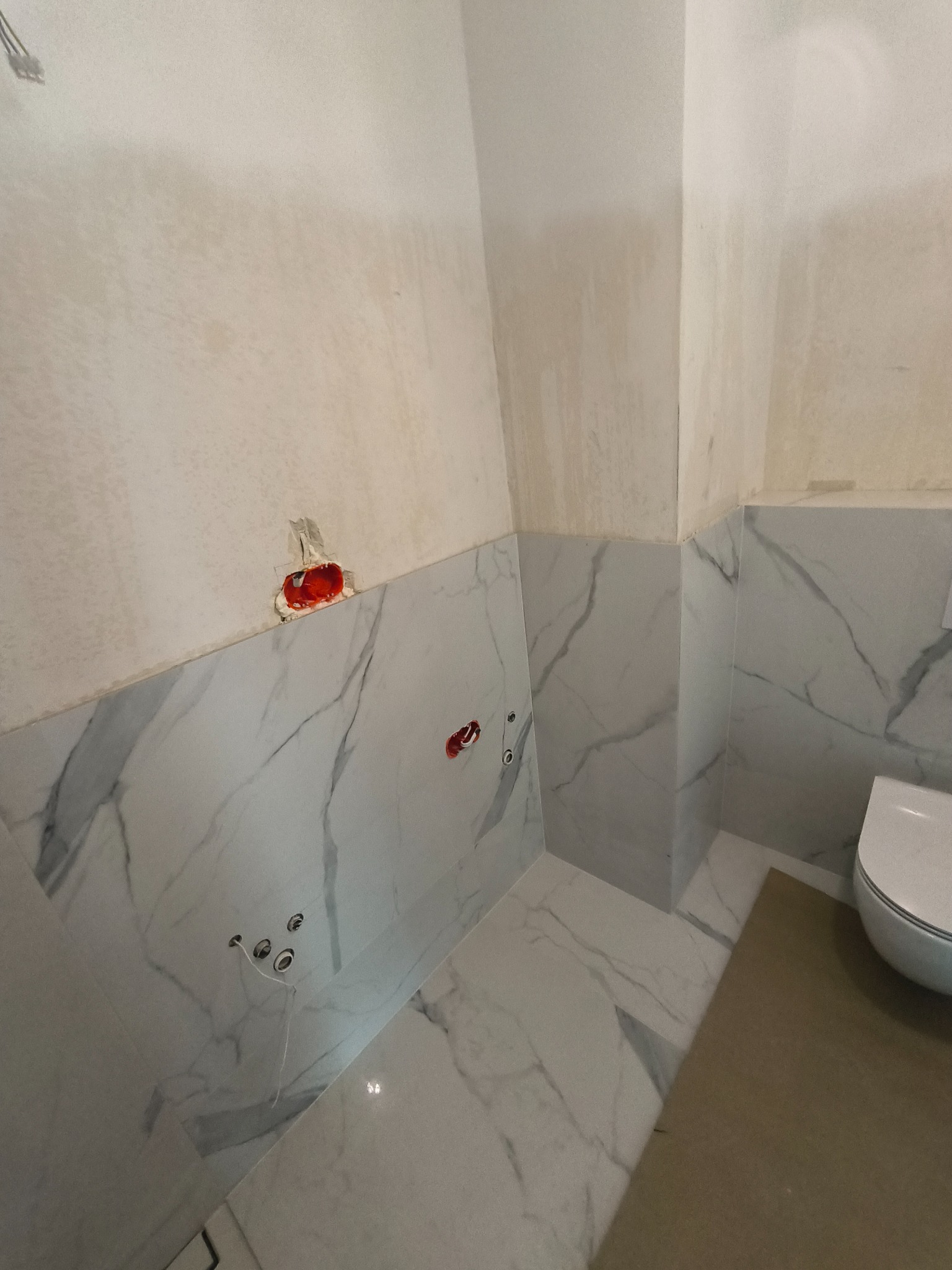 Łazienka w trakcie wykańczania: białe płytki imitujące marmur na ścianach do połowy wysokości, surowy tynk powyżej, widoczne otwory pod instalacje elektryczne i hydrauliczne, zabezpieczona podłoga,...