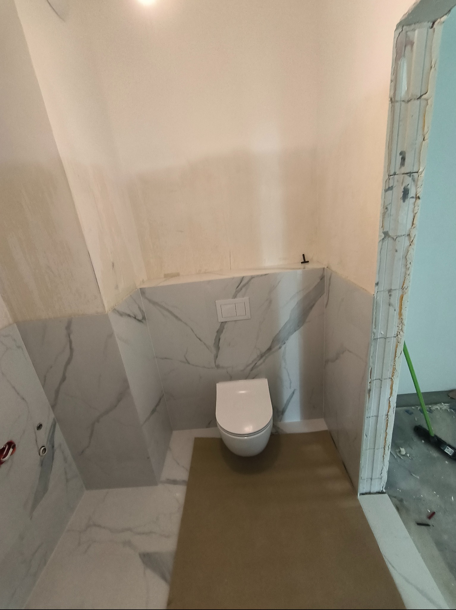 Niedokończona łazienka z częściowo ułożonymi białymi płytkami imitującymi marmur na ścianach, wisząca toaleta i przygotowane podłoże pod posadzkę, widoczny fragment ościeżnicy w stanie surowym.
