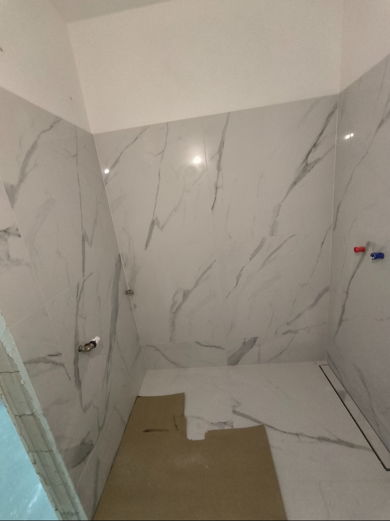 Wnętrze łazienki w trakcie wykańczania, widoczne białe płytki imitujące marmur na ścianach i podłodze, zabezpieczone kartonem. Ujęcie z niskiego kąta, widoczny odpływ liniowy w podłodze i wystające...