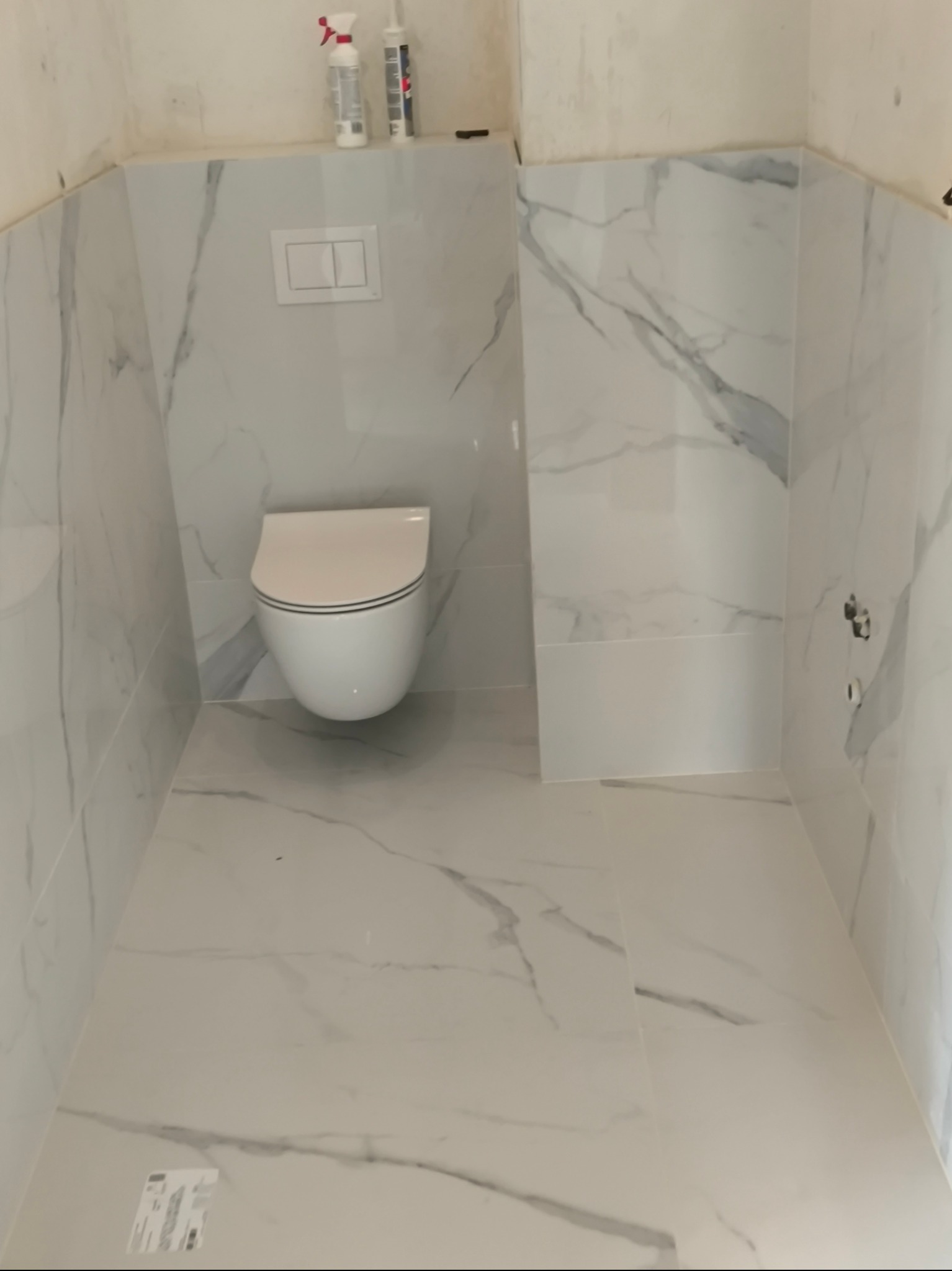 Nowoczesna, minimalistyczna łazienka z białymi płytkami imitującymi marmur, wisząca toaleta i zabudowany system spłukiwania, widoczne niedoskonałości wykończenia na górze ściany.