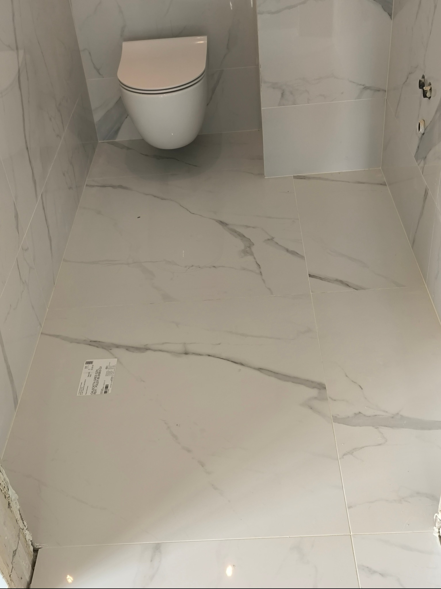 Wykończona, jasna łazienka z podwieszaną toaletą i płytkami imitującymi marmur na ścianach i podłodze, widoczna naklejka producenta płytek.