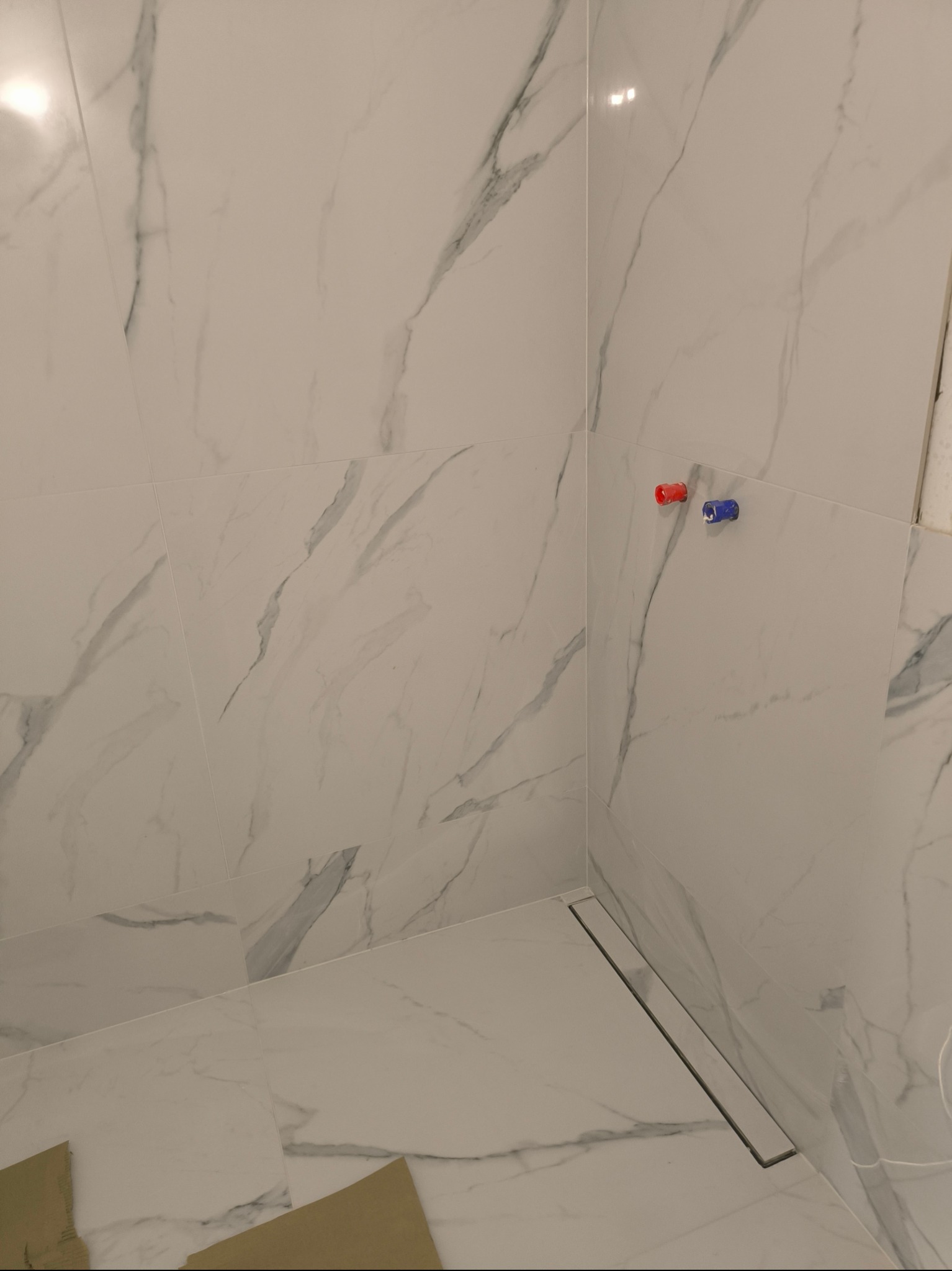 Wykończona ściana i podłoga w łazience z białych płytek imitujących marmur, widoczny odpływ liniowy i zaślepione zawory wody w kolorach czerwonym i niebieskim.