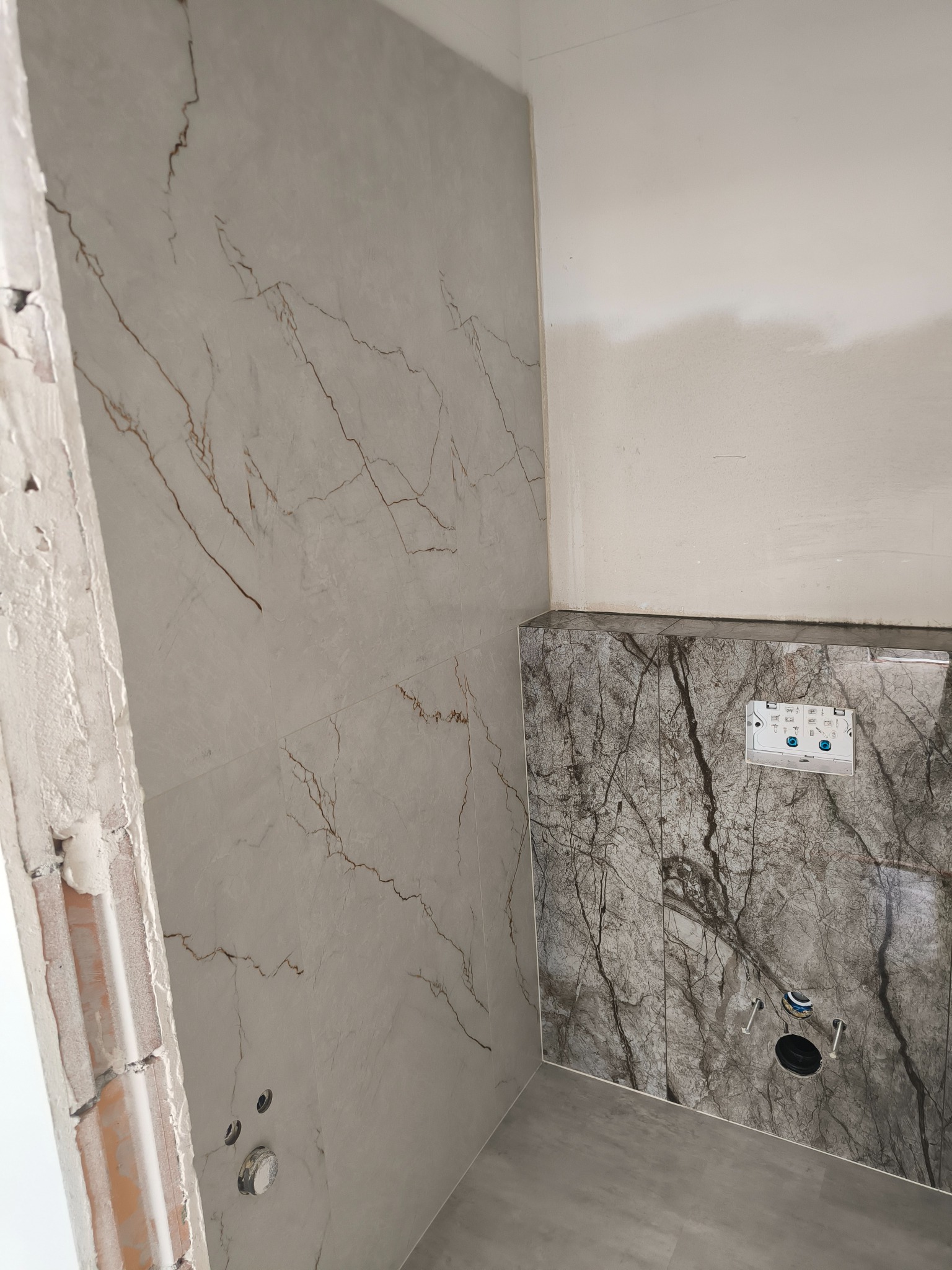 Montaż płytek imitujących marmur w łazience, widoczne otwory na armaturę i stelaż podtynkowy, ściana częściowo otynkowana, surowy stan wykończenia przy futrynie.