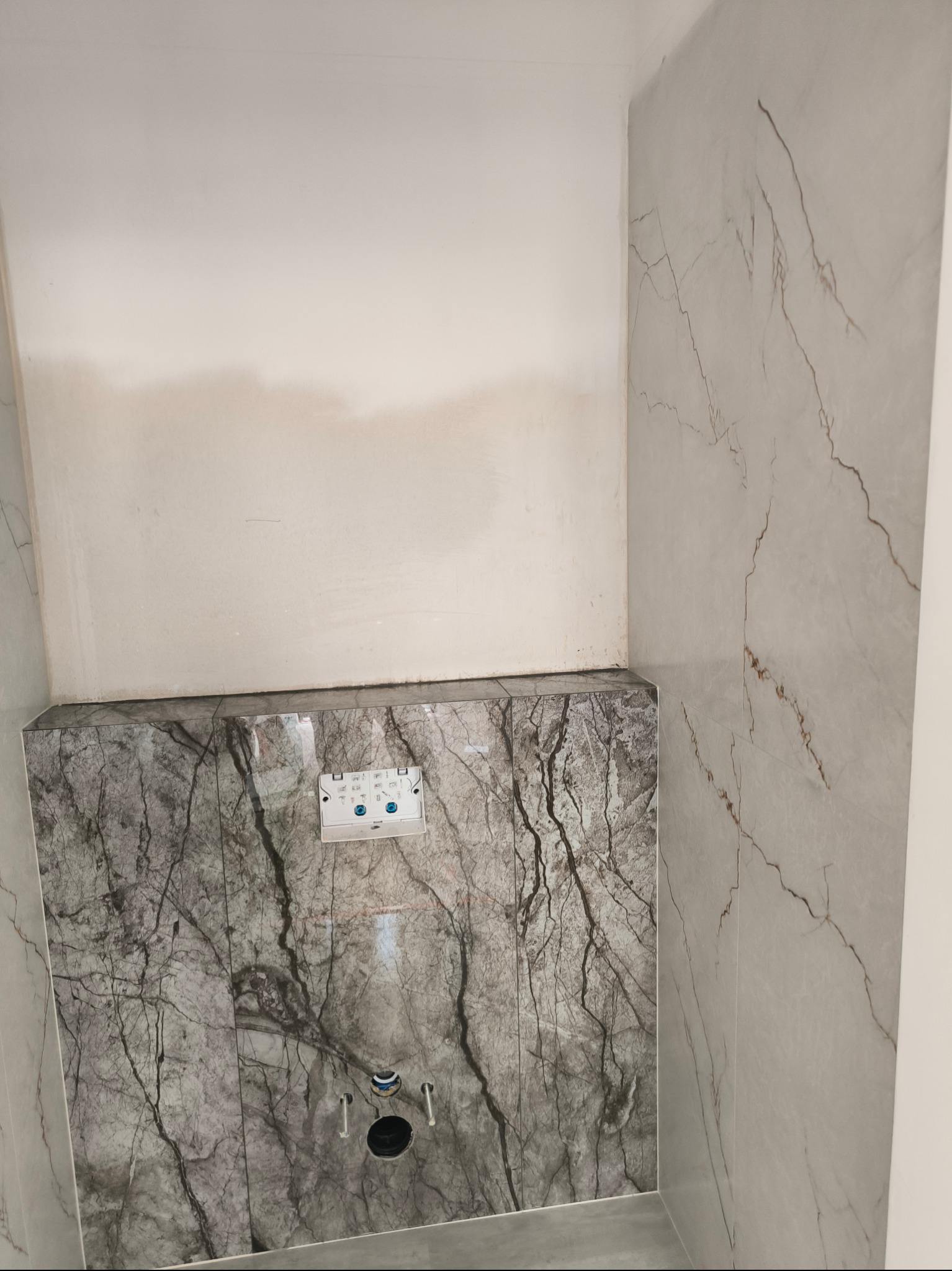 Montaż stelaża podtynkowego WC z obudową z płytek imitujących marmur, widoczne przyłącza wodne i odpływowe, ściany wykończone płytkami imitującymi marmur o jaśniejszym odcieniu, surowy tynk...