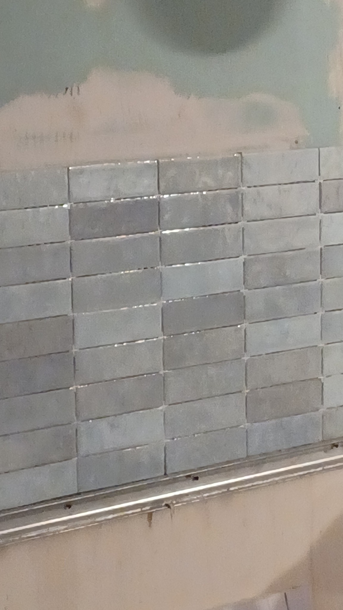 Ściana w trakcie układania podłużnych, szarych płytek, widoczny profil wykończeniowy przy dolnej krawędzi, ślady zaprawy na ścianie powyżej.