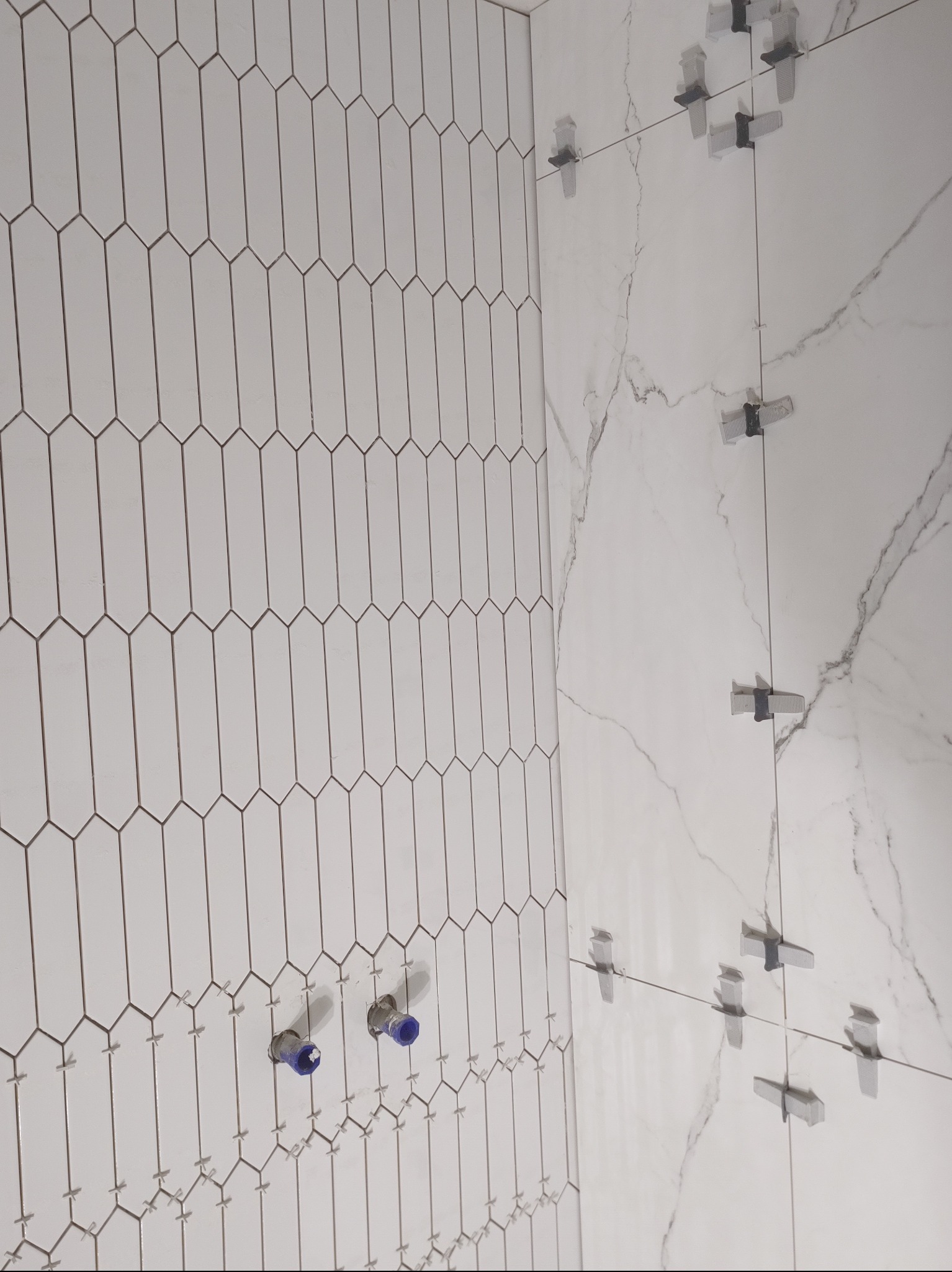 Montaż białych, podłużnych płytek w kształcie migdałów na ścianie, z widocznymi niebieskimi zaślepkami na rurach i systemem poziomowania płytek.