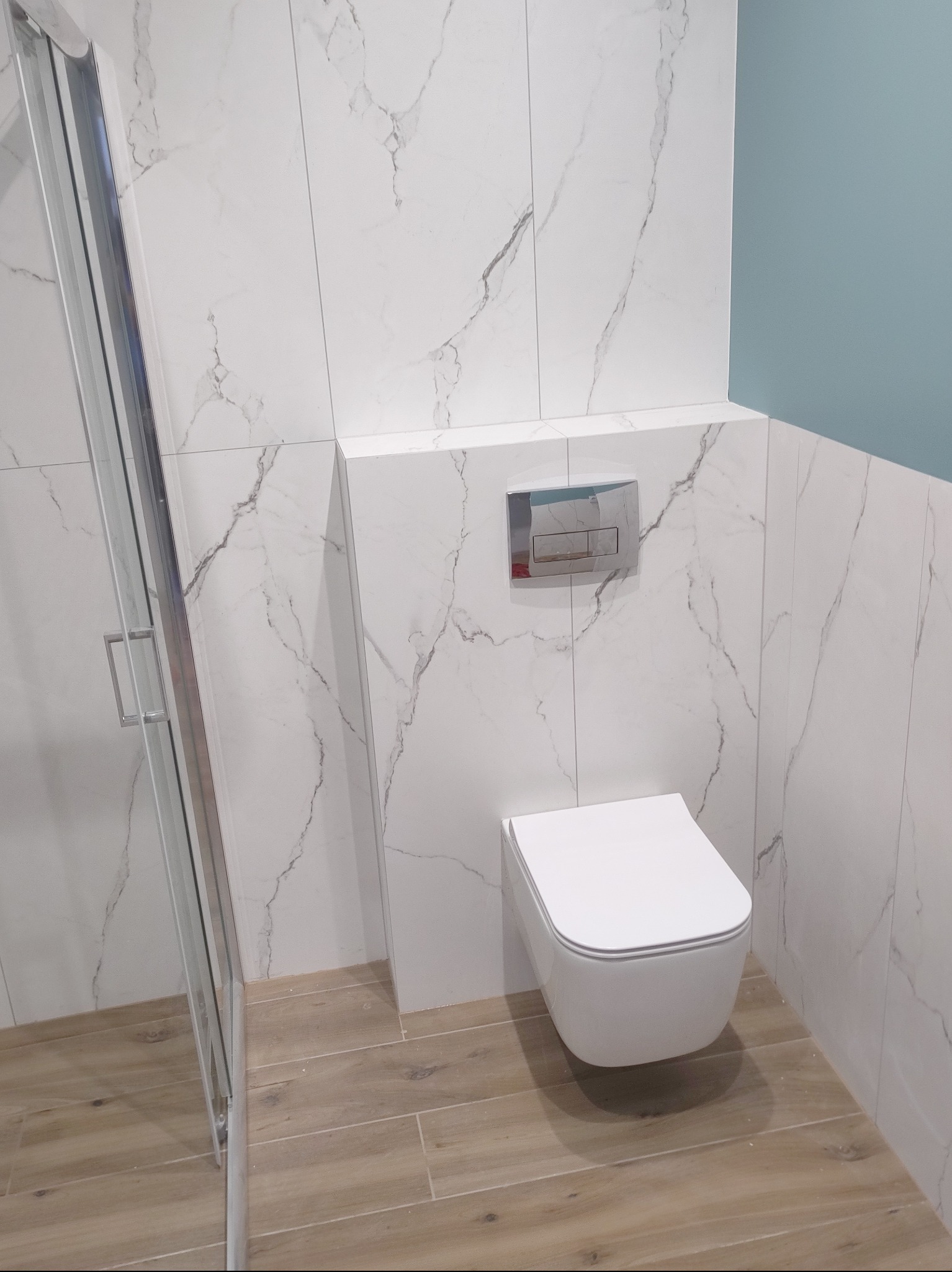 Nowoczesna łazienka z białymi płytkami imitującymi marmur, wisząca toaleta i drewnopodobna podłoga, kabina prysznicowa ze srebrnymi profilami, ściana pomalowana na jasny odcień niebieskiego.