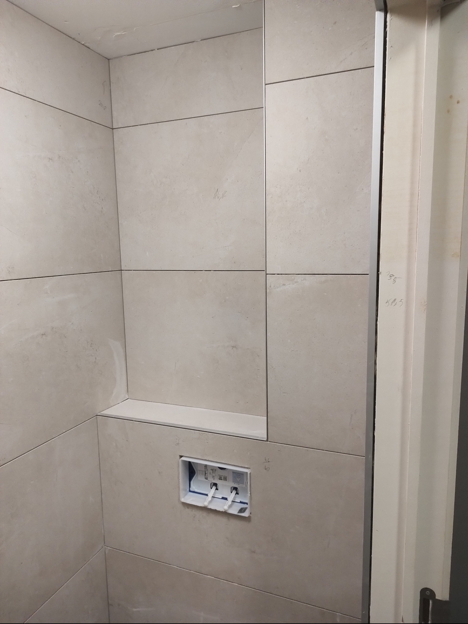 Ściana łazienki wyłożona beżowymi płytkami ceramicznymi, wnęka z półką i zamontowany stelaż podtynkowy WC, widoczne notatki na płytkach i ościeżnicy.