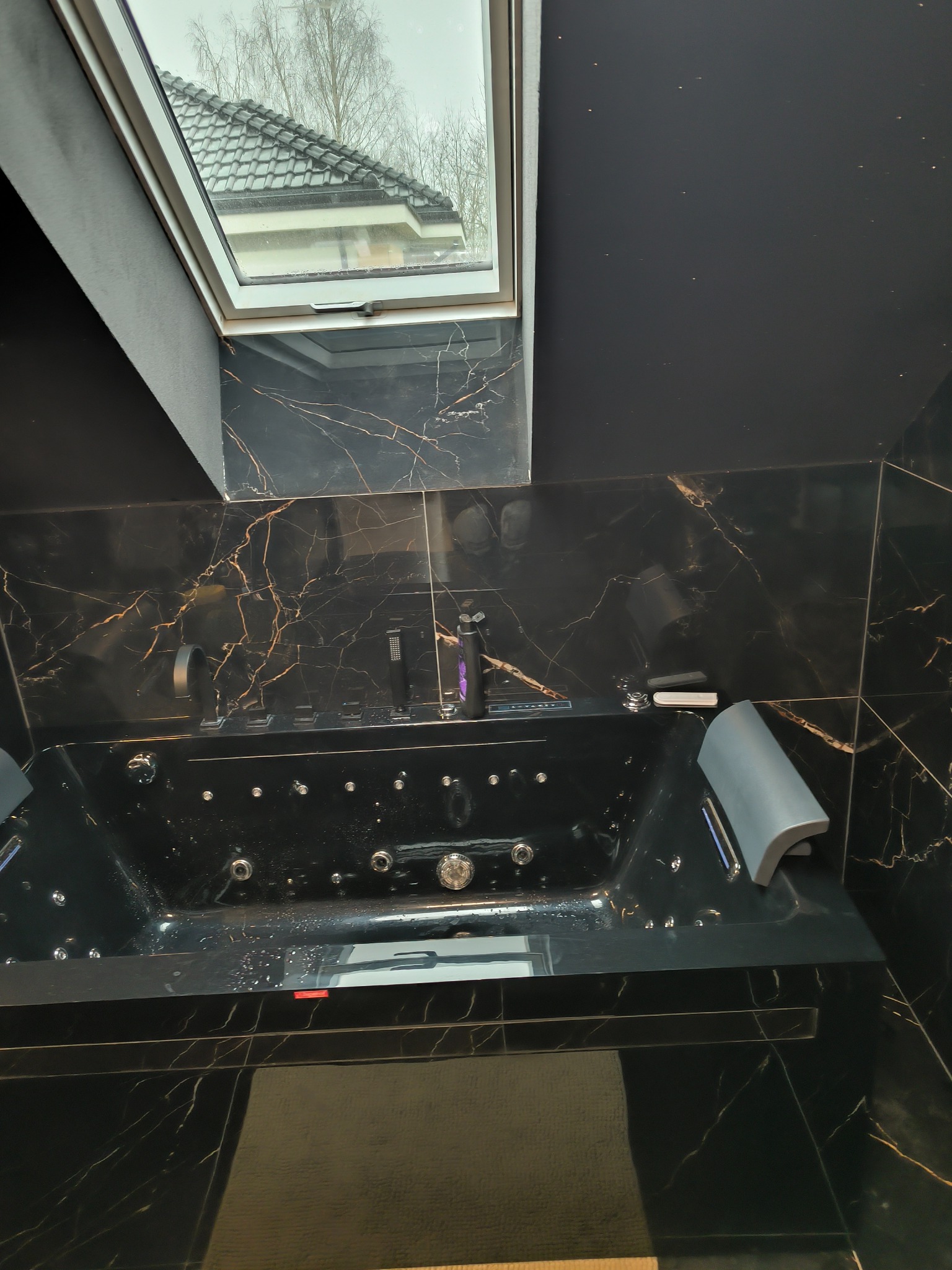 Czarna wanna z hydromasażem pod oknem dachowym, wykończona płytkami imitującymi marmur ze złotymi żyłami. Nowoczesna łazienka po remoncie.