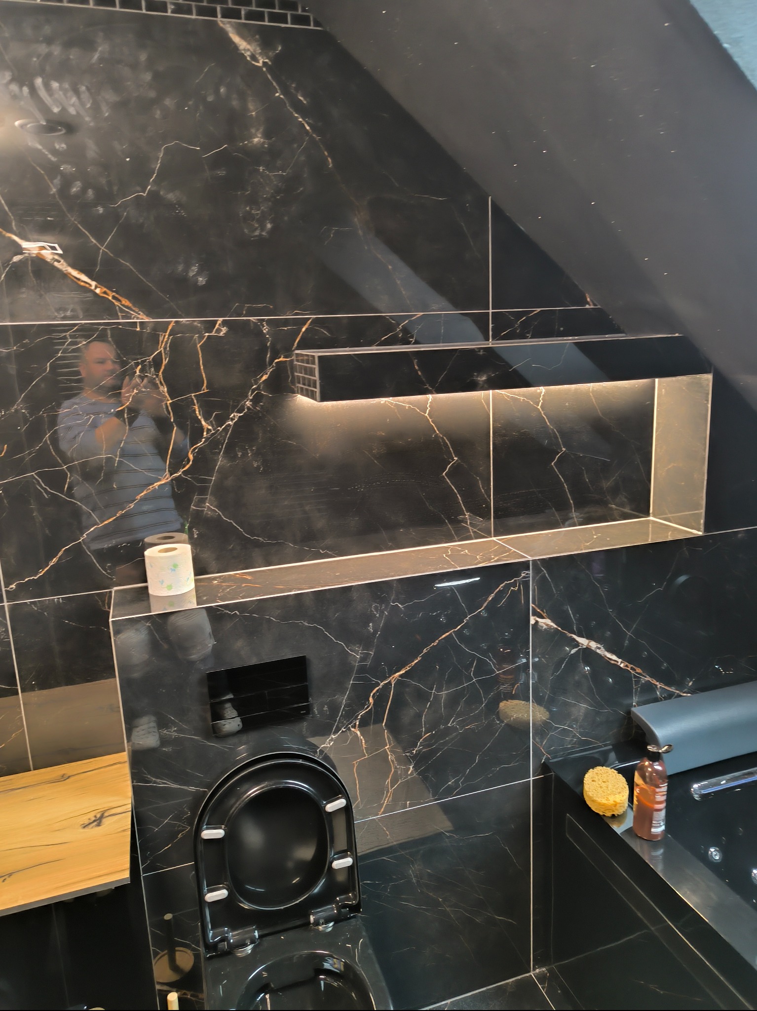 Nowoczesna łazienka z czarnymi płytkami imitującymi marmur, podświetlana wnęka w ścianie, wisząca toaleta i fragment drewnianego blatu. Widoczne akcesoria łazienkowe.