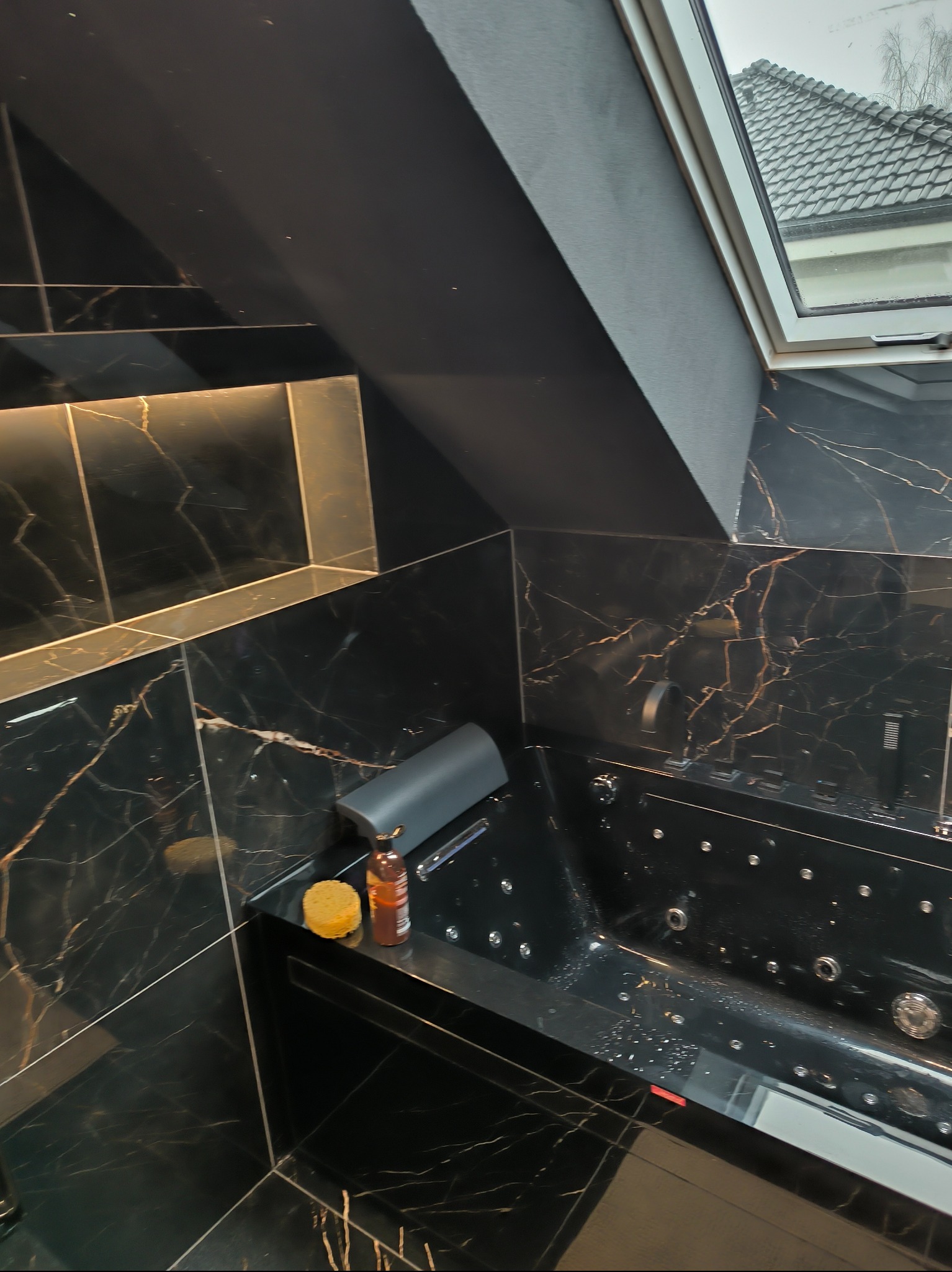 Czarna łazienka z marmurowymi płytkami i wanną z hydromasażem pod oknem dachowym. Na blacie gąbka i butelka. Oświetlenie LED podkreśla luksusowy charakter.