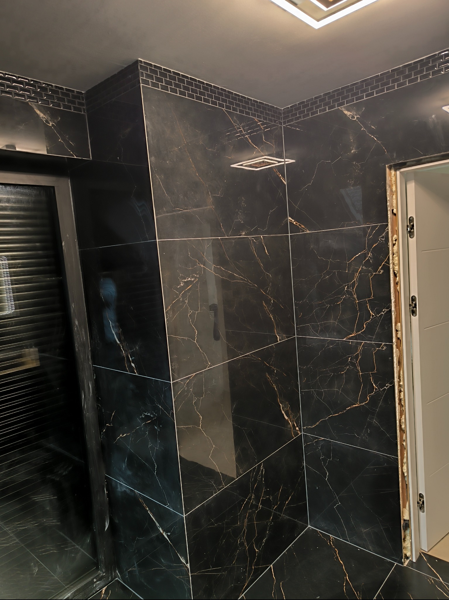 Czarne, marmurowe płytki z dekoracyjnym paskiem mozaiki w wykończonej łazience. Widoczny fragment kabiny prysznicowej i oświetlenie sufitowe.