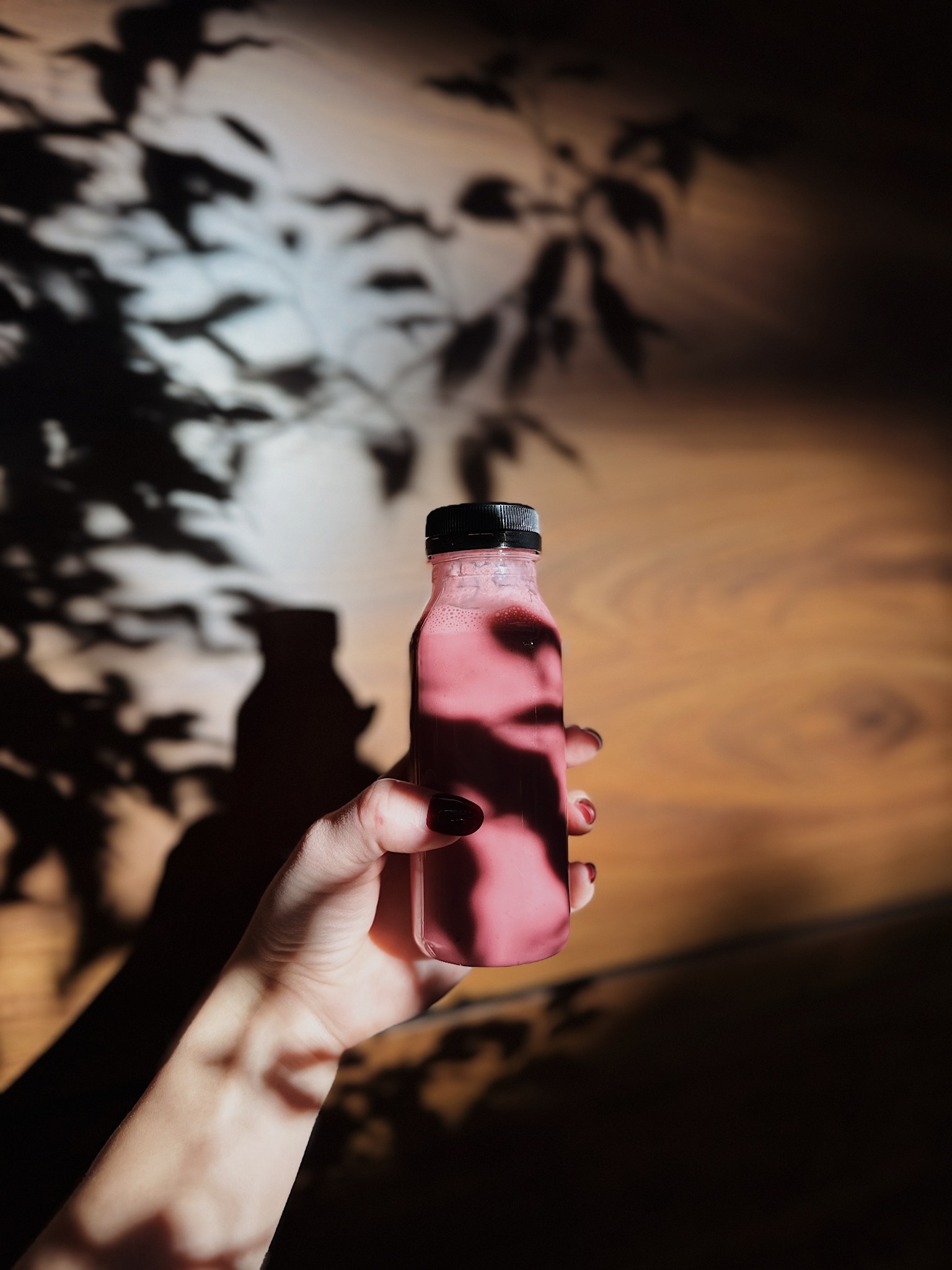 Dłoń trzyma butelkę różowego smoothie na tle drewnianej ściany z cieniem liści. Kontrastowe oświetlenie podkreśla naturalny skład napoju.