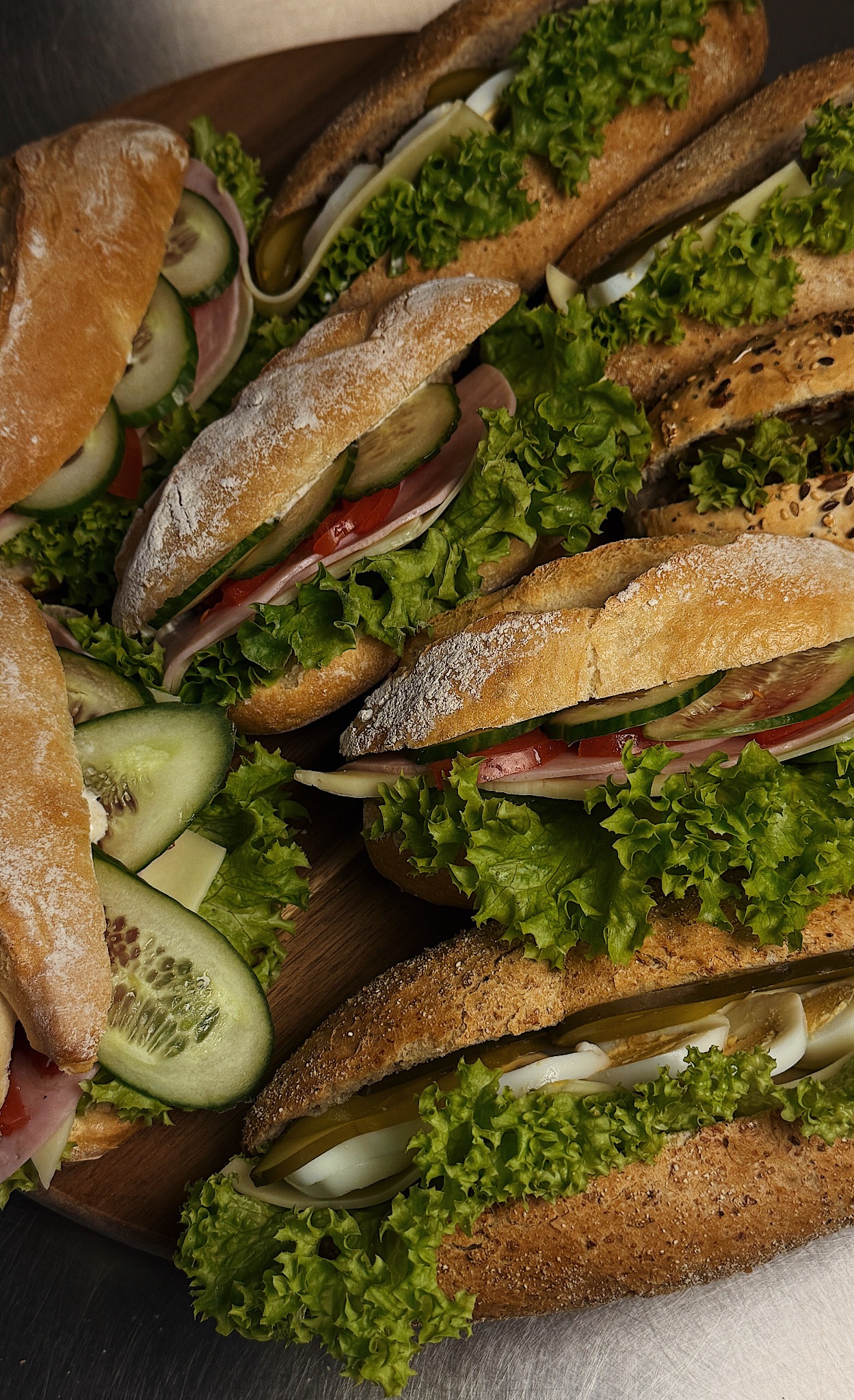 Różnorodne kanapki na drewnianej desce: jasne i ciemne pieczywo, szynka, ser, jajko, ogórek, pomidor i sałata, tworzące apetyczną kompozycję.