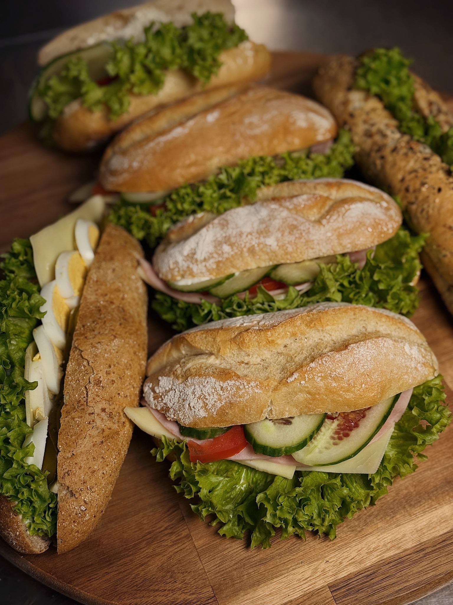 Różnorodne kanapki na drewnianej desce: bagietki z szynką, serem, jajkiem, pomidorem, ogórkiem i sałatą, idealne na catering.