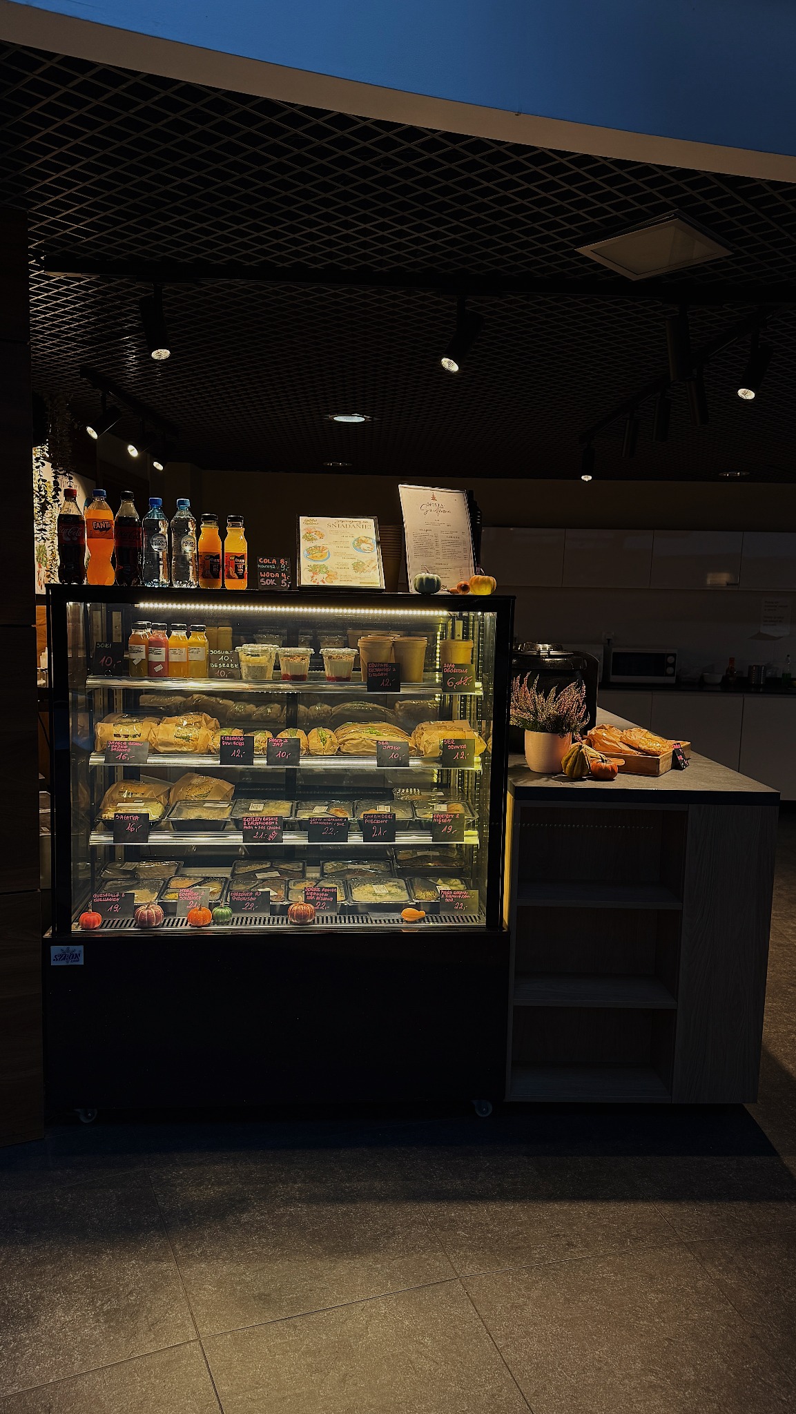 Chłodnicza witryna w bistro z napojami, deserami i daniami gotowymi, menu na górze, dekoracje z dyni. Widok na ladę i zaplecze.
