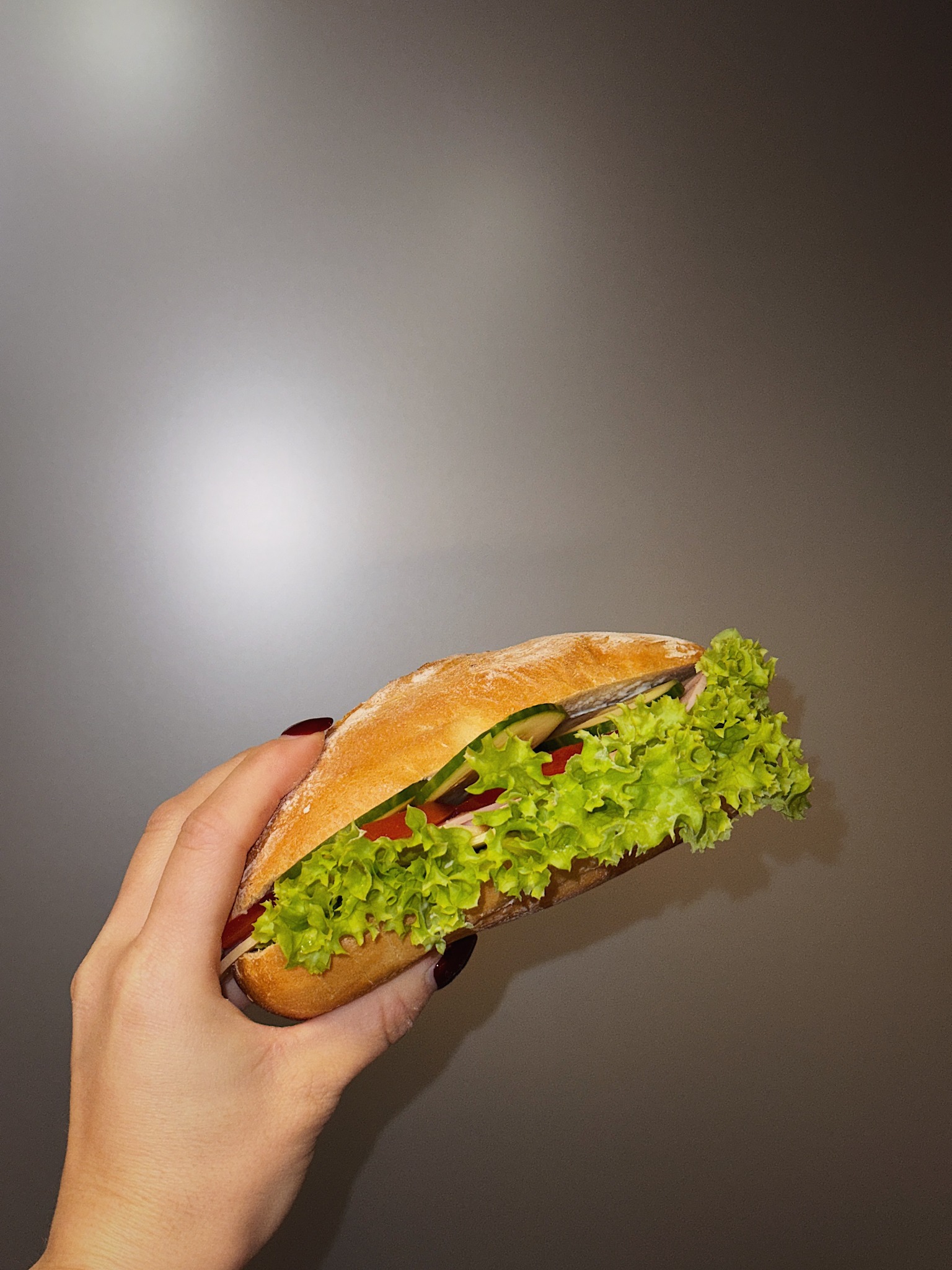 Świeża kanapka ciabatta z zieloną sałatą, pomidorem i ogórkiem, trzymana w dłoni z ciemnym manicure na tle szarego tła.