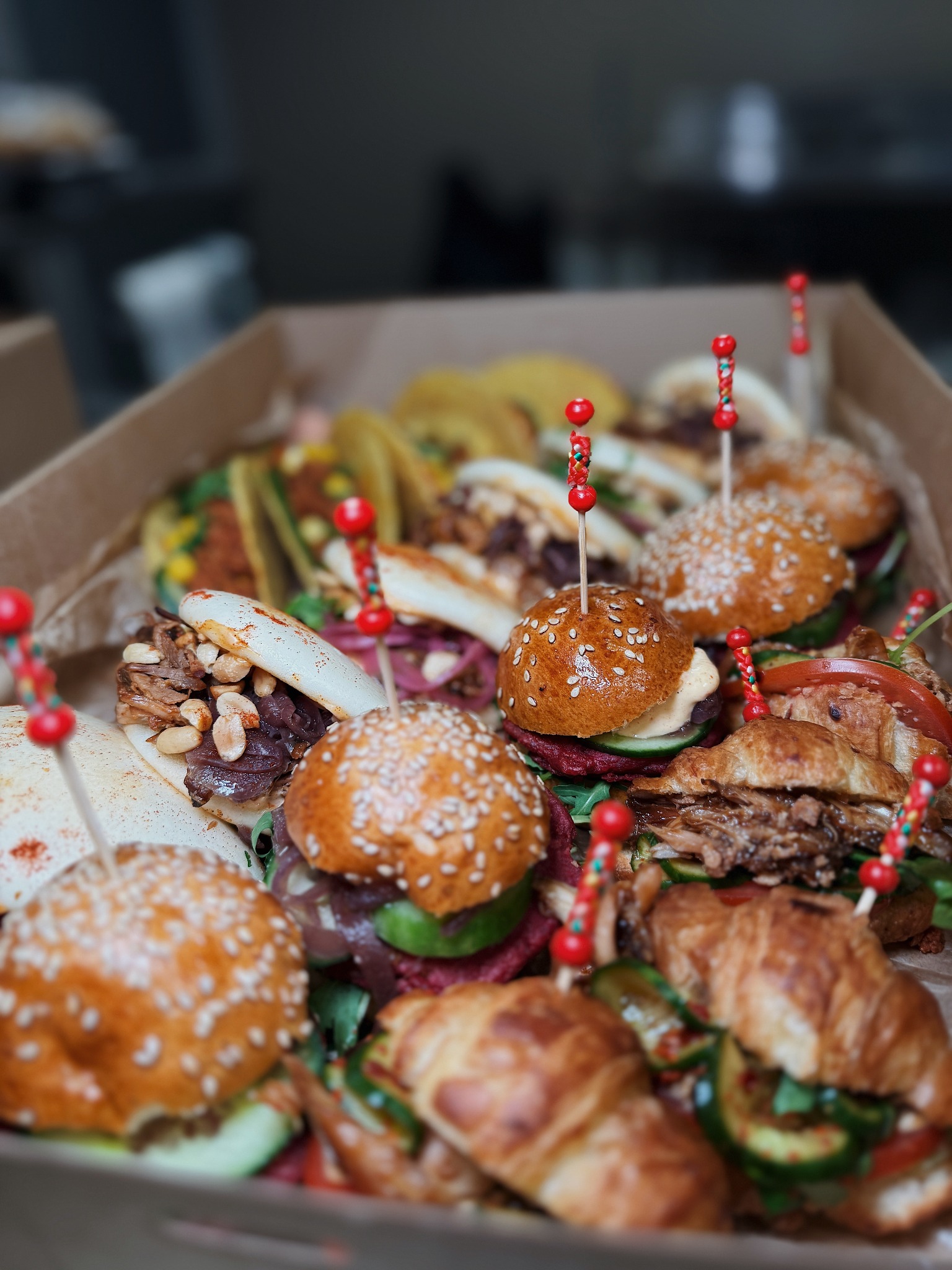 Różnorodny box finger food: mini burgery z sezamem, tacos i bao z mięsem, ozdobione kolorowymi wykałaczkami, w tekturowym pudełku cateringowym.