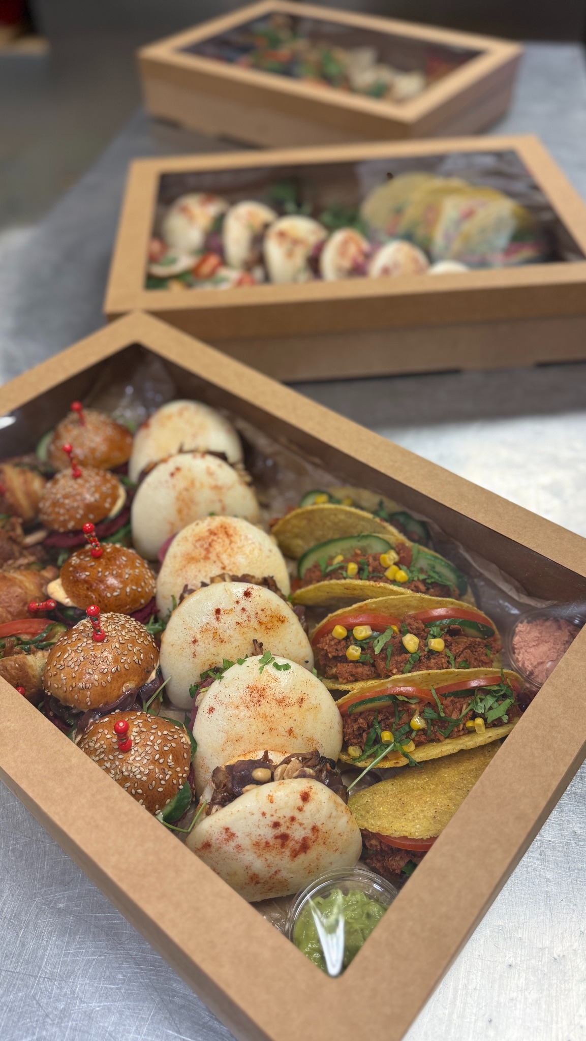Box finger food: mini burgery z czerwonymi wykałaczkami, bao z posypką, wegańskie tacos z kukurydzą i dipy w przezroczystych pojemnikach. Drugi box w tle.