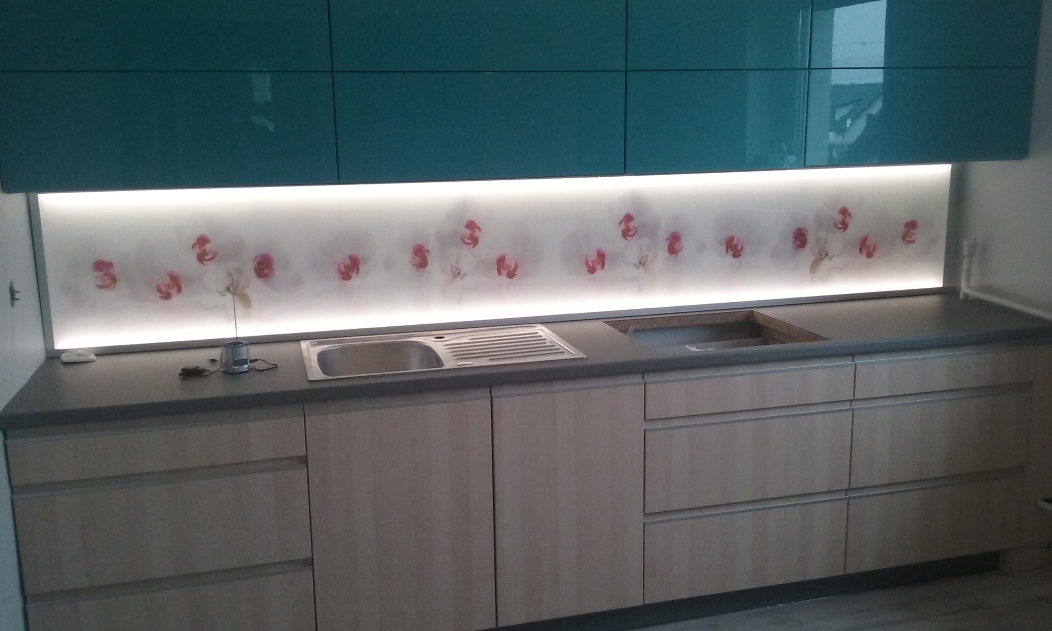 Nowoczesna zabudowa kuchenna z jasnymi, drewnopodobnymi frontami szafek, szarym blatem i turkusowymi szafkami górnymi. Ściana nad blatem zdobiona podświetlanym panelem z motywem białych orchidei.