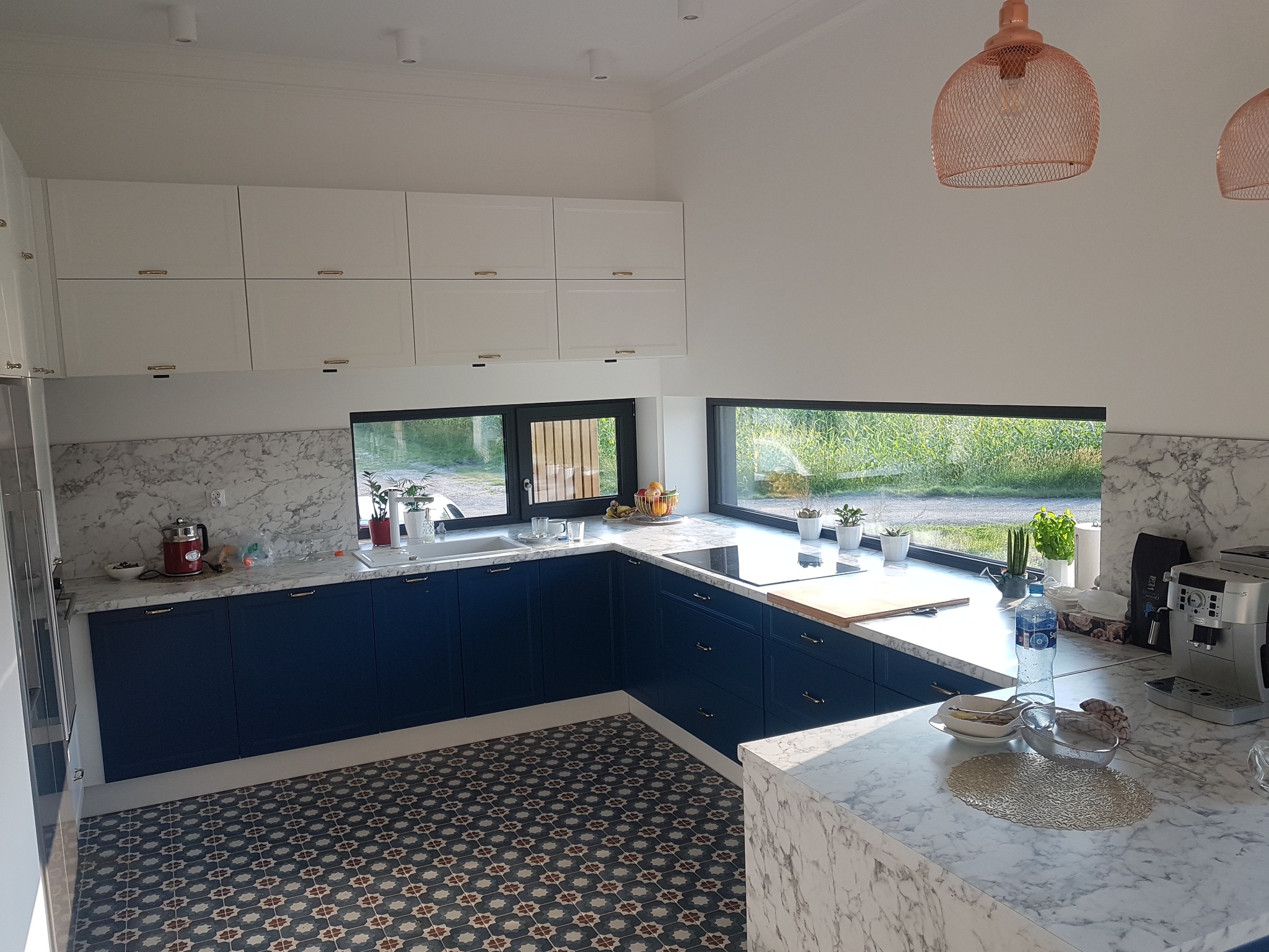 Nowoczesna kuchnia z granatowymi szafkami dolnymi i białymi górnymi, marmurowy blat i backsplash, okna z widokiem na zieleń, wzorzysta podłoga, oświetlenie w stylu industrialnym.