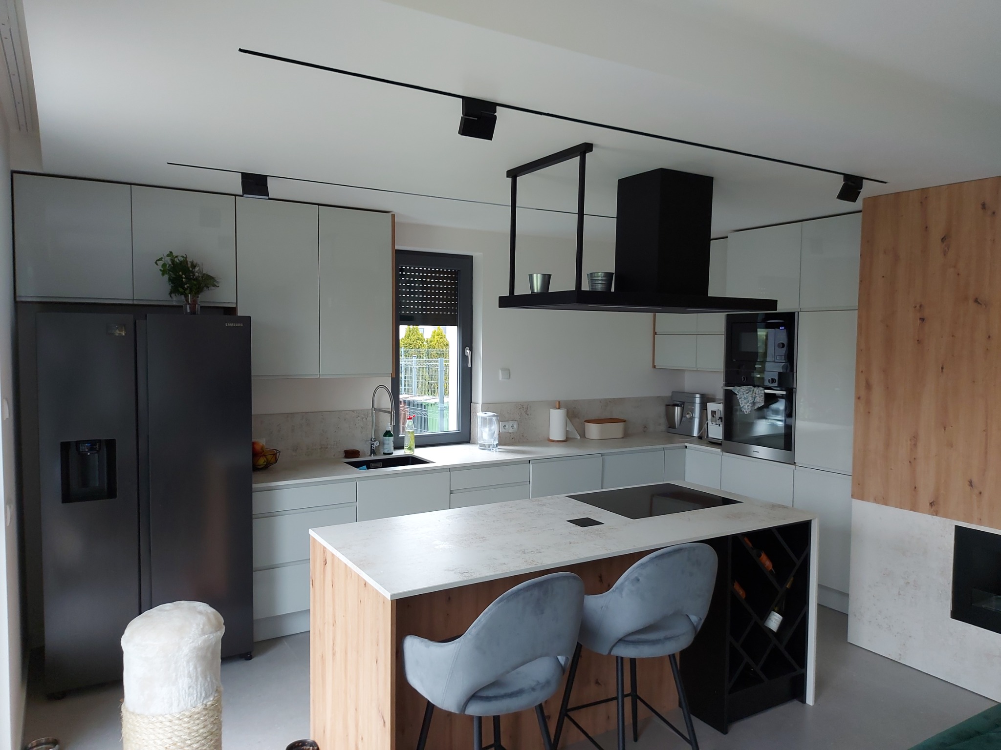 Nowoczesna kuchnia z wyspą, białymi szafkami, czarną lodówką i okapem oraz drewnianymi akcentami, oświetlona listwami LED.