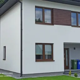 Nowoczesny dom jednorodzinny z oknami PCV w kolorze drewna, z naklejką firmy ochroniarskiej na jednym z okien, widoczne pojemniki do segregacji odpadów przed wejściem.