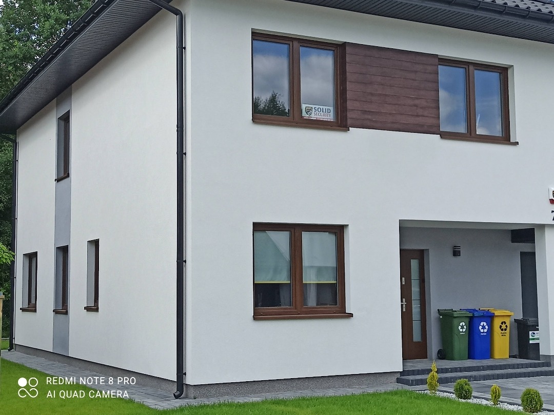 Nowoczesny dom jednorodzinny z oknami PCV w kolorze drewna, z naklejką firmy ochroniarskiej na jednym z okien, widoczne pojemniki do segregacji odpadów przed wejściem.