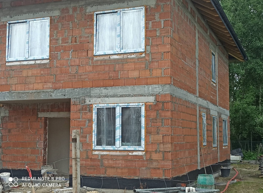 Surowy budynek z czerwonej cegły z zamontowanymi białymi oknami zabezpieczonymi folią ochronną, widoczne materiały budowlane i fragment dachu.