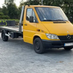 Żółta laweta Mercedes-Benz Sprinter z platformą załadunkową i zielonym elementem konstrukcyjnym na platformie, zaparkowana na szarym bruku w słoneczny dzień.