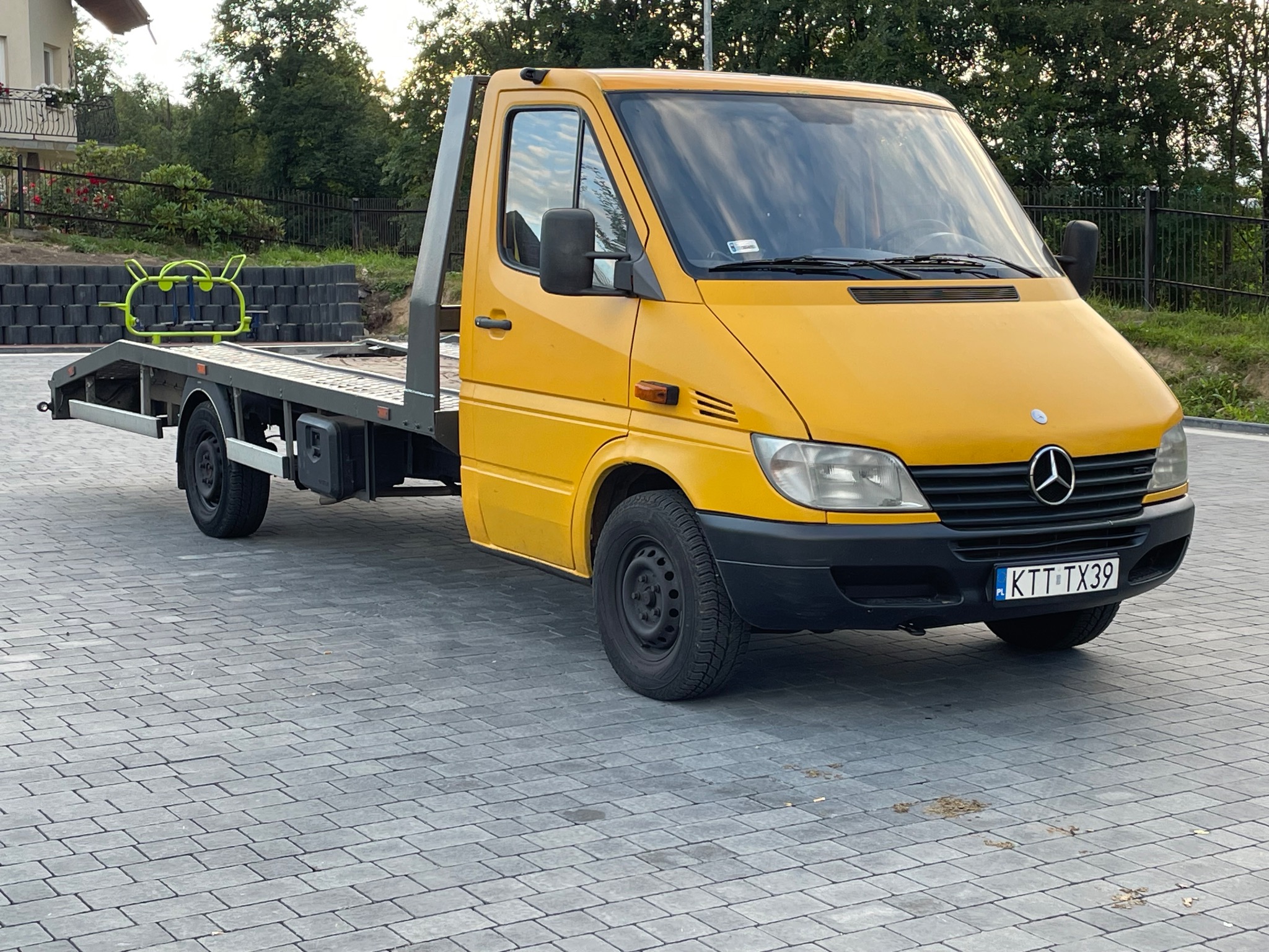 Żółta laweta Mercedes-Benz Sprinter z platformą załadunkową i zielonym elementem konstrukcyjnym na platformie, zaparkowana na szarym bruku w słoneczny dzień.