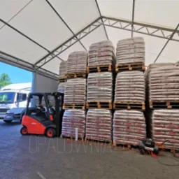 Magazyn pelletu: wózek widłowy Linde obok palet z pelletem poltarex.pl pod zadaszeniem, w tle biała ciężarówka Mercedes