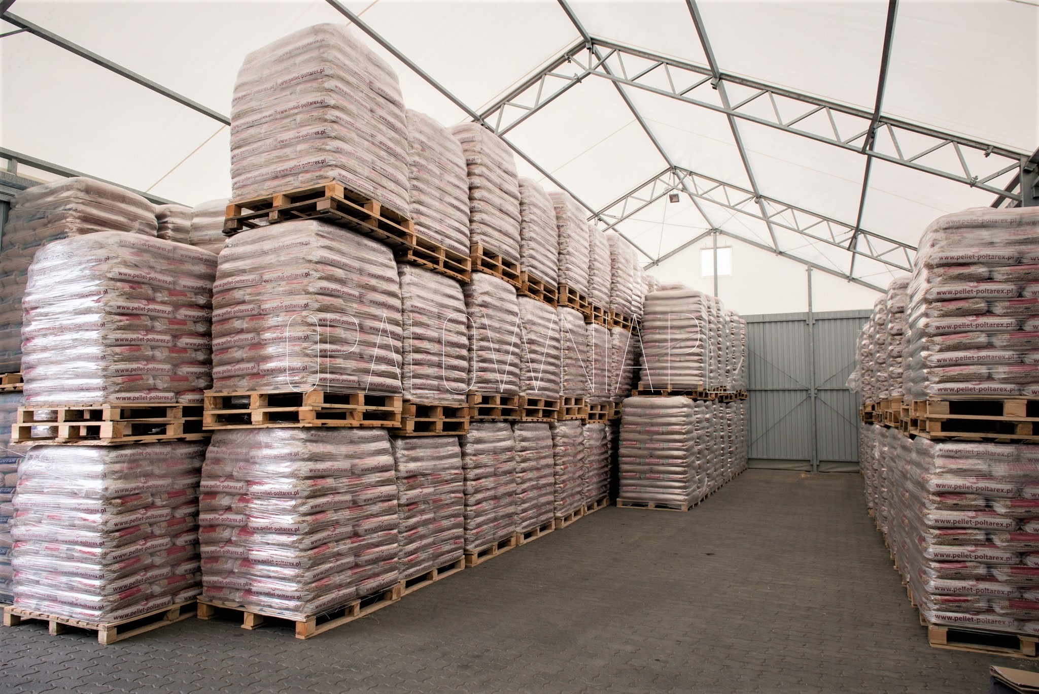 Magazyn pelletu: stosy worków z pelletem ułożone na paletach pod zadaszeniem magazynowym, widoczne metalowe elementy konstrukcji dachu.